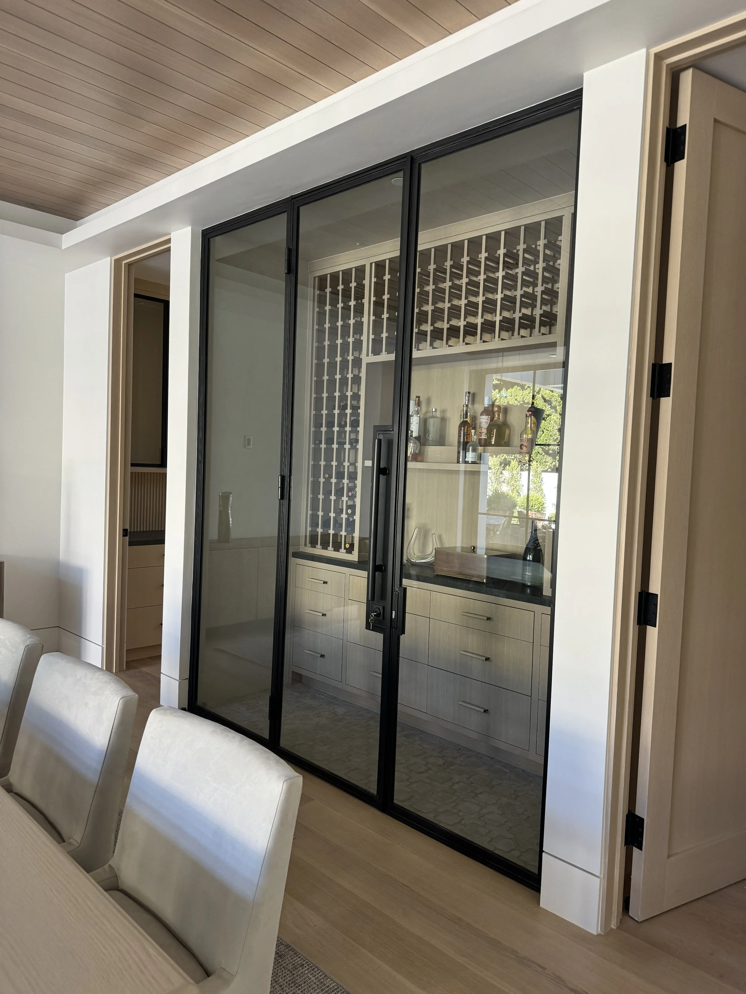 Living room with a glass partition door, wooden ceiling, light-colored furniture, and a built-in cabinet with bottles and glassware behind the glass.