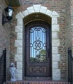 Front door with decorative glass and wrought iron design, surrounded by brick wall and stone archway, with lantern-style light fixture to the left.
