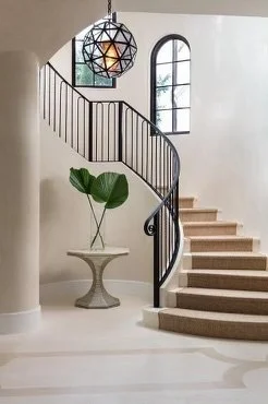 Interior of a modern stairway with a black railing, a large window, a decorative plant on a pedestal, and a geometric pendant light hanging from the ceiling.