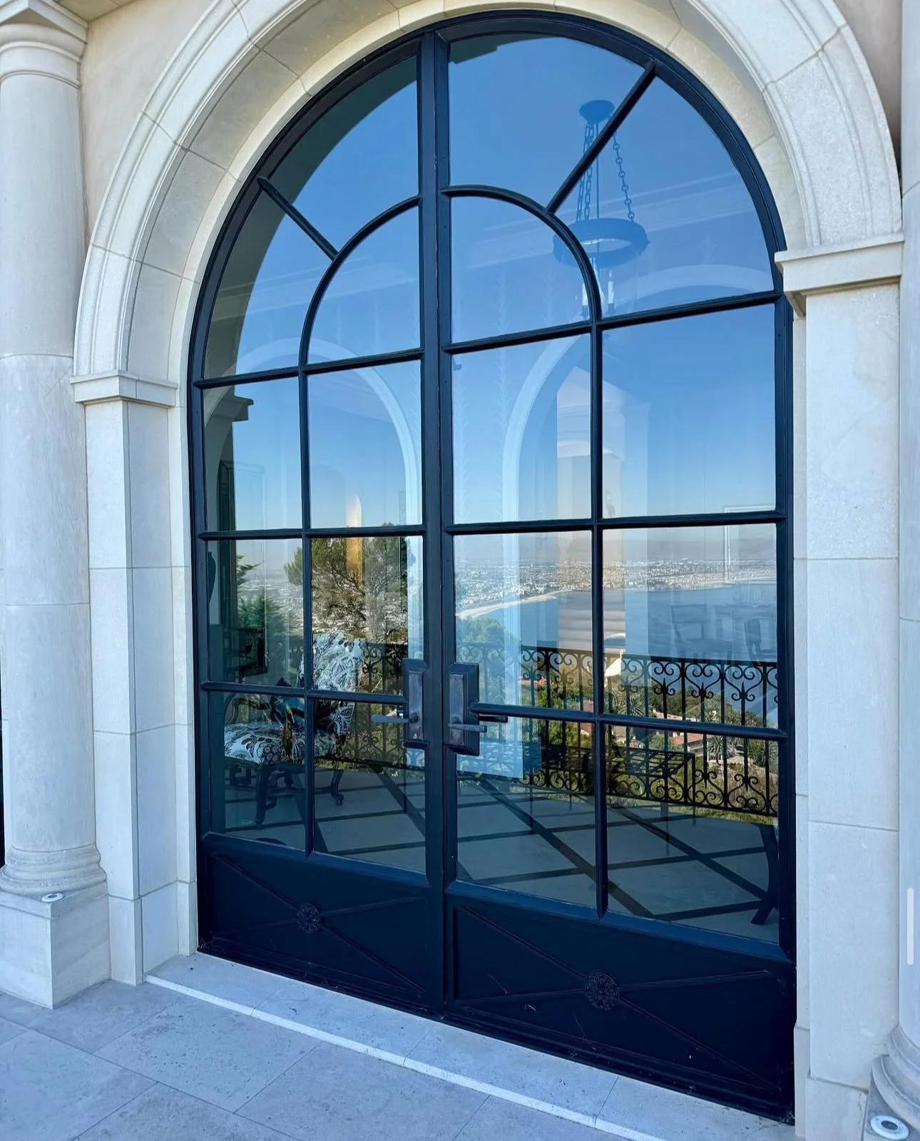 Large decorative window with a door, reflecting the scenery outside, including a body of water, a city skyline, and a chandelier inside.