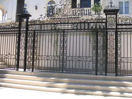 Ornate black wrought iron gate with staircase in front, leading to a building with white exterior and blue accents.