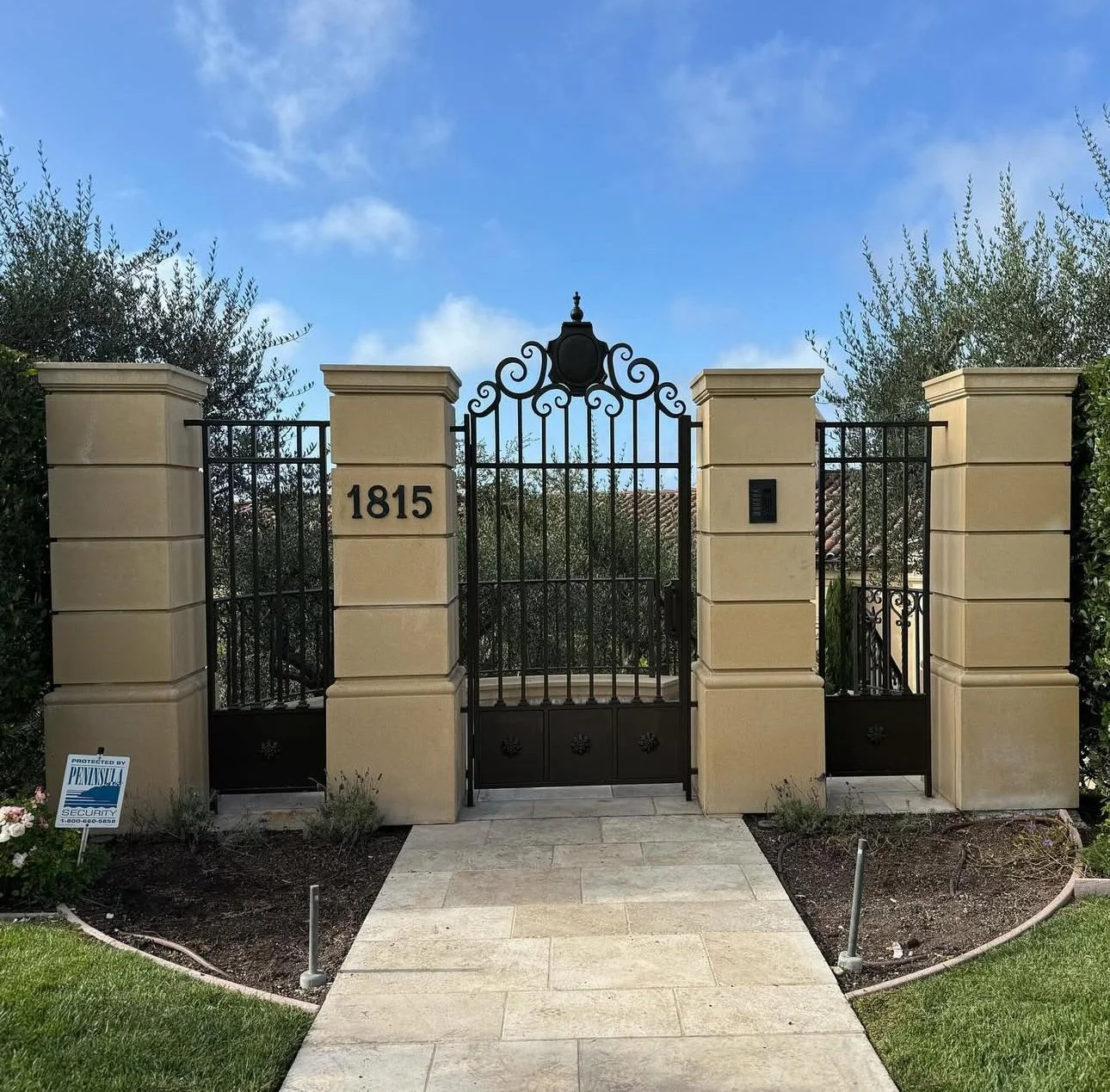 A black wrought iron gate with decorative swirl designs, supported by beige stone pillars, leading to a residential property with the house number 1815 displayed on the left pillar. A pathway leads to the gate, with green grass and flowerbeds on eith