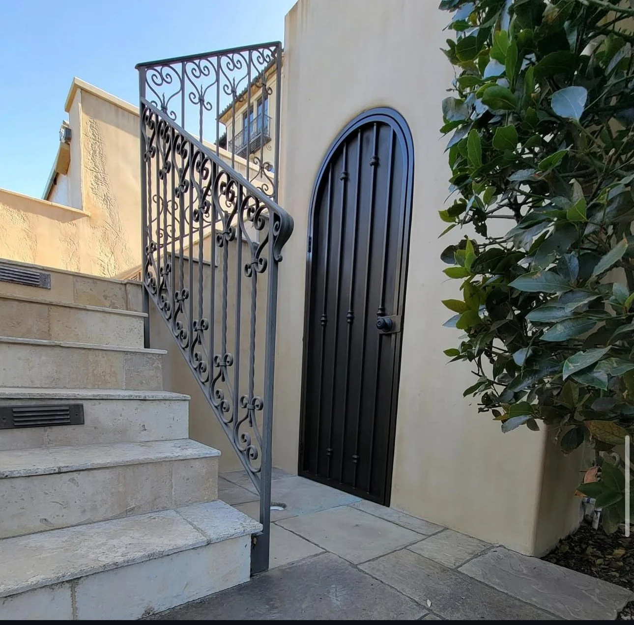 Stone stairs with iron railing leading to a black arched door on a beige stucco wall, with green shrubbery to the right and a blue sky above.
