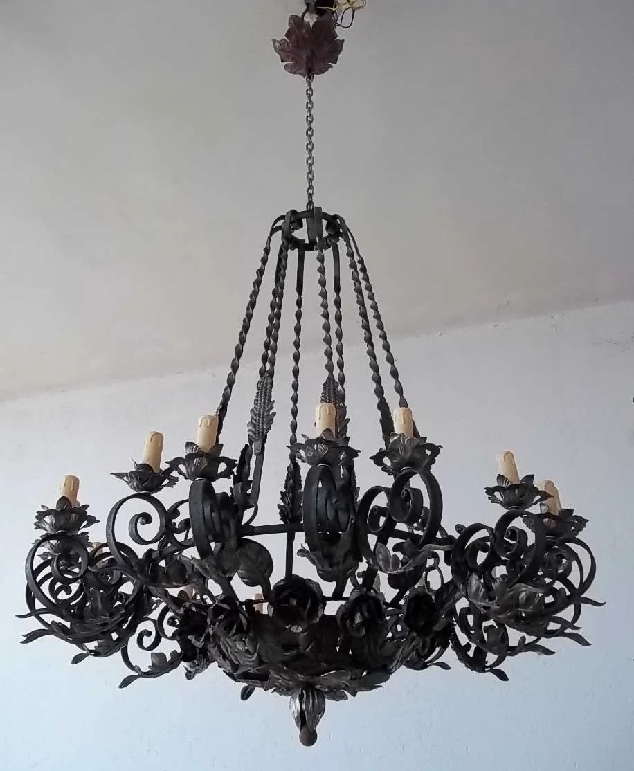 Black metal chandelier with intricate floral and scrollwork design and multiple candle-like light fixtures hanging from chains.