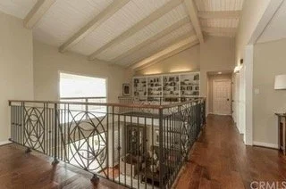 Interior view of a house with a railing around a staircase, hardwood floors, a window letting in natural light, and a hallway leading further inside.