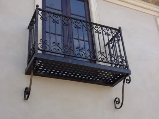 Small decorative black metal balcony with ornate railing, attached to a beige building with a large purple window.
