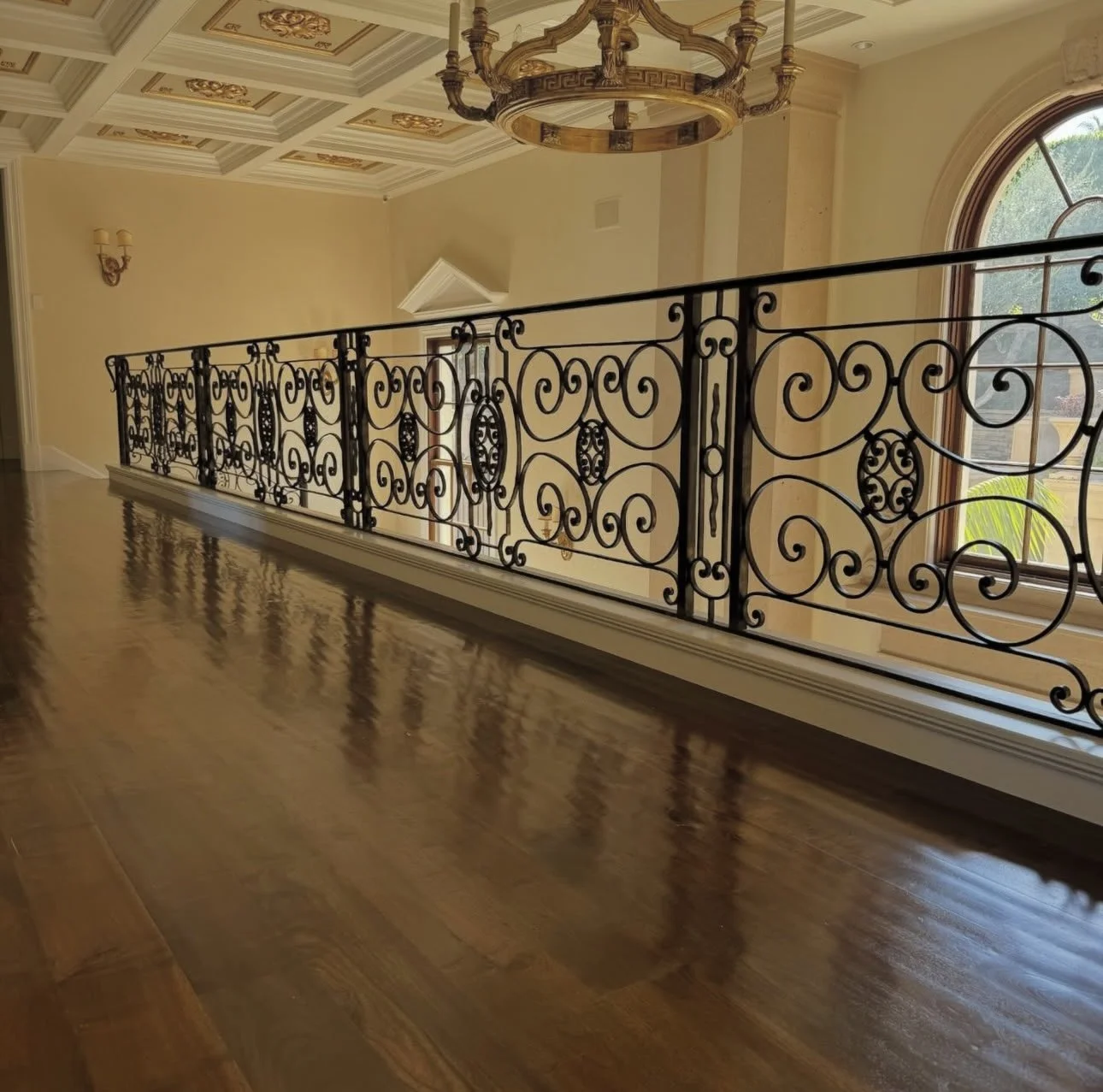 Interior of a house with ornate iron railing, high ceiling with decorative panels, chandelier, and a large window with a view outside.