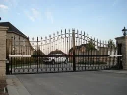 A black metal gate with decorative spikes, closed, in front of an estate with stone walls and buildings beyond.