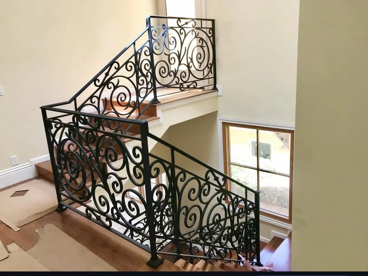 Interior view of a staircase with ornate black wrought iron railing, wooden steps, and large window letting in natural light.
