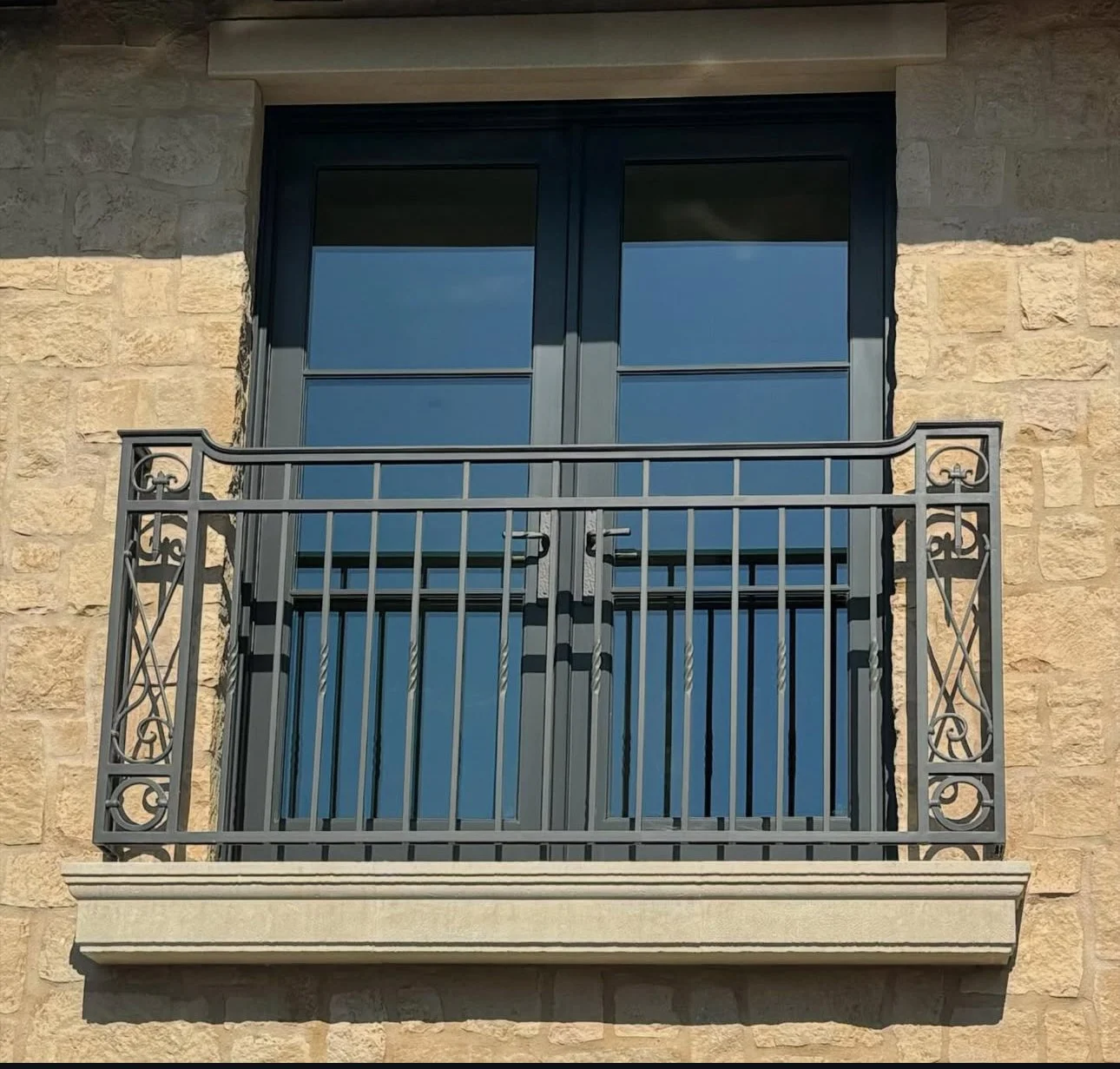 Image of a small balcony with a black metal railing and glass double doors, attached to a beige stone building.
