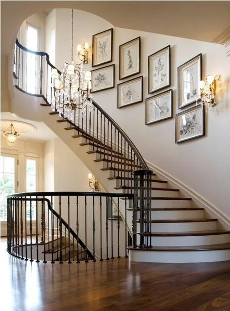 Elegant staircase with wooden steps and black iron railing in a well-lit hallway, decorated with framed botanical artwork and chandeliers.