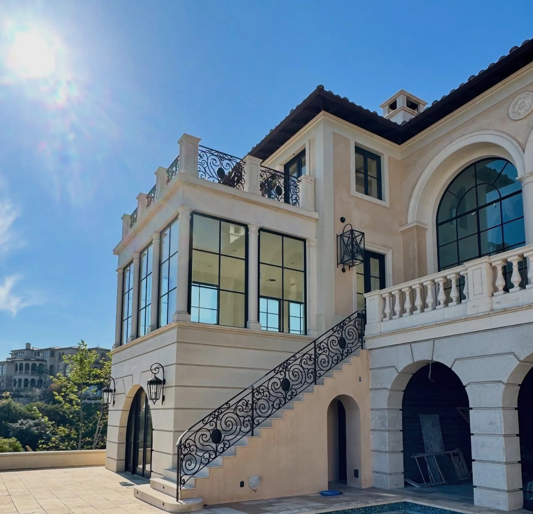 Luxury house with large glass windows, ornate wrought iron staircase, and Mediterranean architectural style, under a bright blue sky.