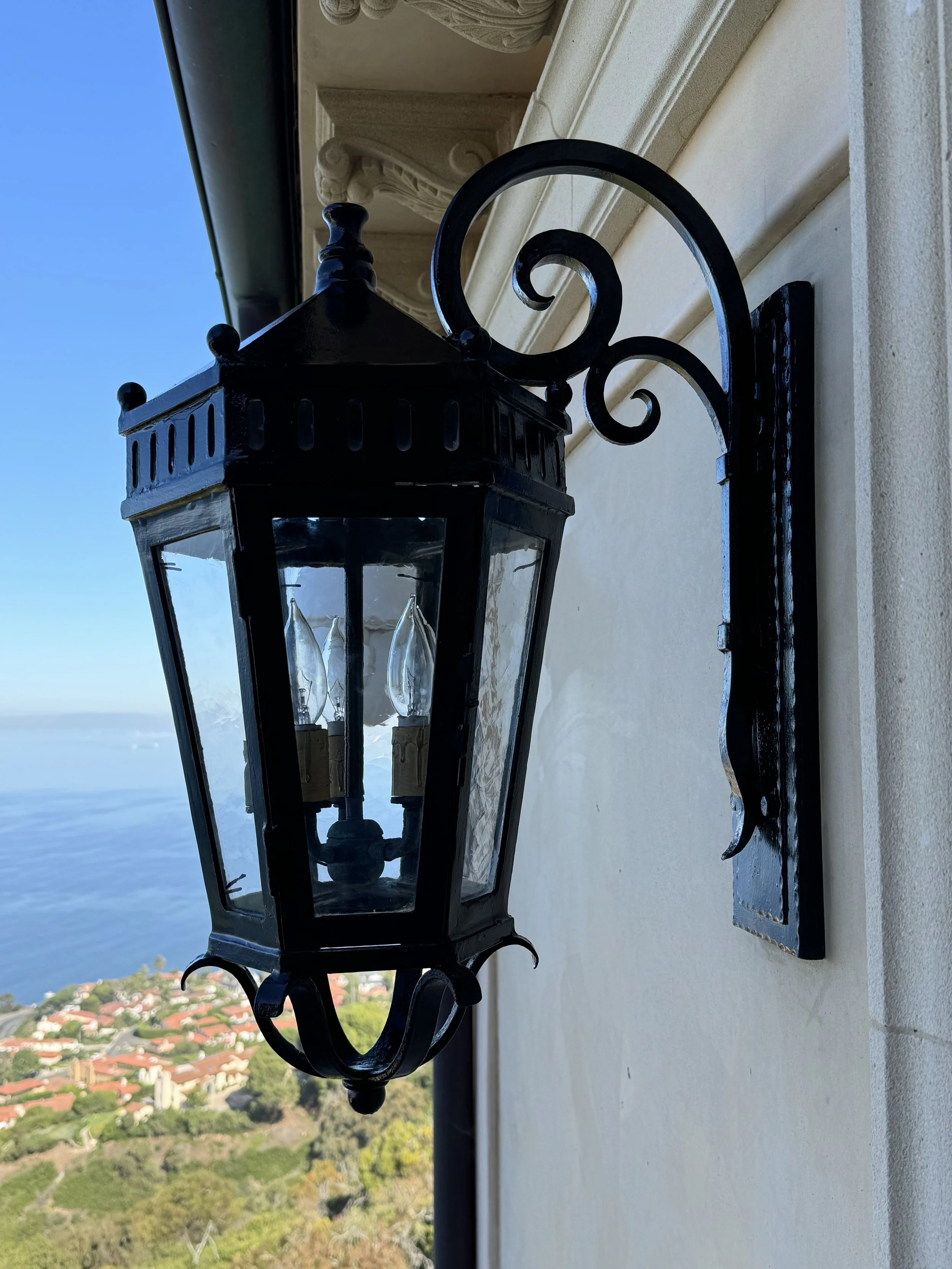 Close-up of a black outdoor lantern attached to a beige wall with a scenic coastal view in the background.