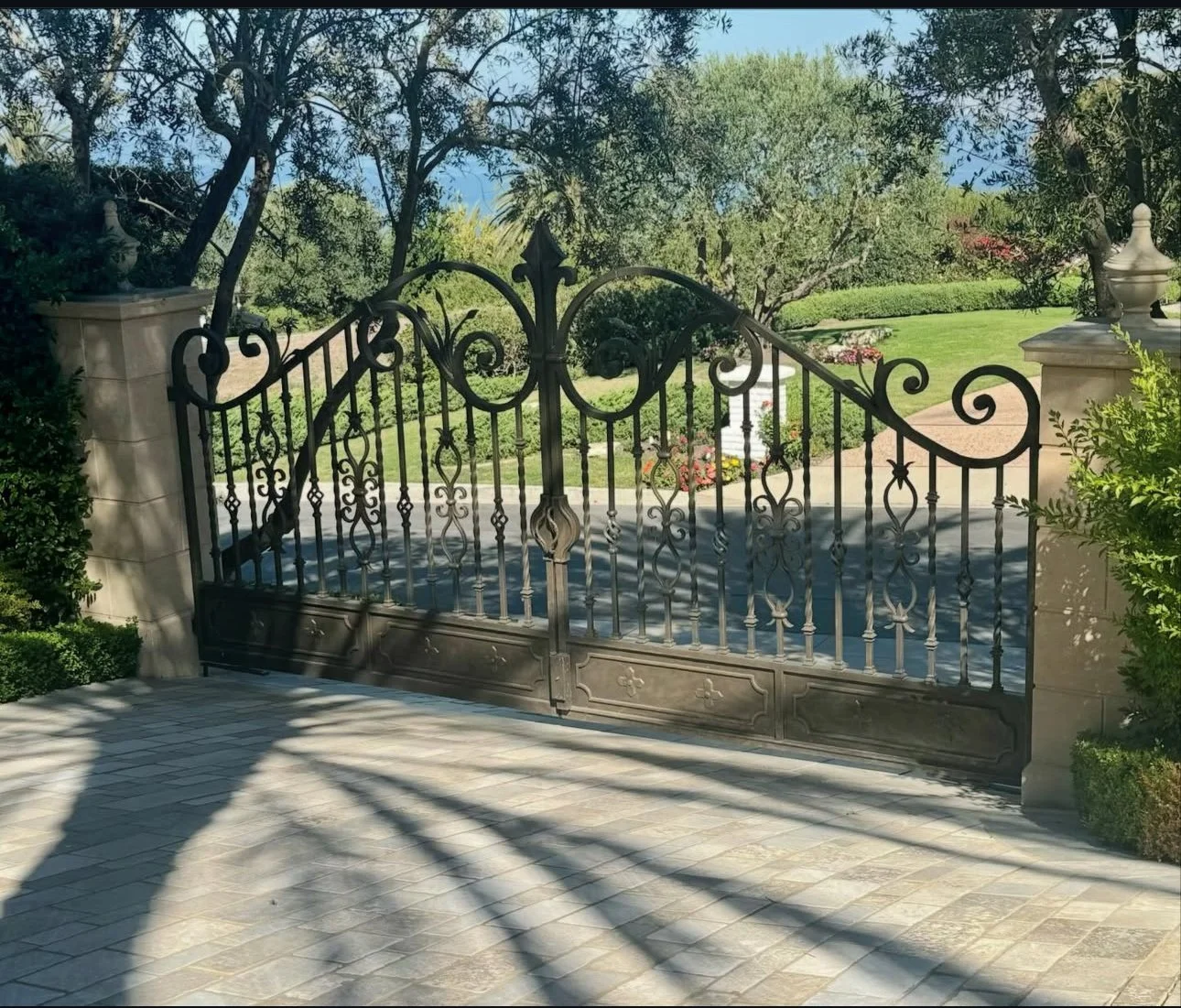Black wrought iron gate with decorative scrollwork, flanked by stone pillars, leading to a garden with lush green grass, trees, and flower beds, on a sunny day.