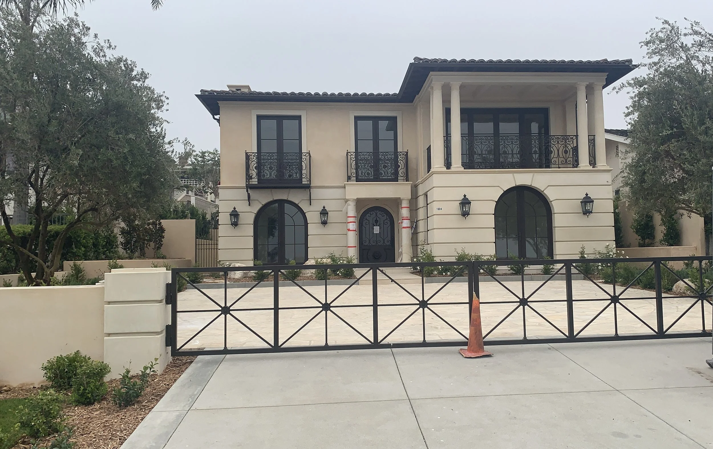 A two-story modern house with beige walls, black window frames, and a black door. The house features small balconies with decorative black railings and outdoor wall lanterns. A driveway with a parked traffic cone is in front of the house, and there a