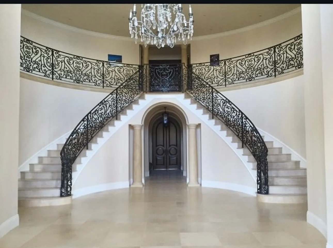 Elegant foyer with curved staircase featuring ornate black wrought iron railings, a chandelier hanging from the ceiling, and a dark double door archway at the base of the stairs.