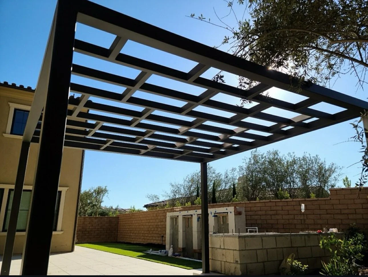 Backyard patio with a modern pergola structure and a brick wall, with nearby trees and a house visible.