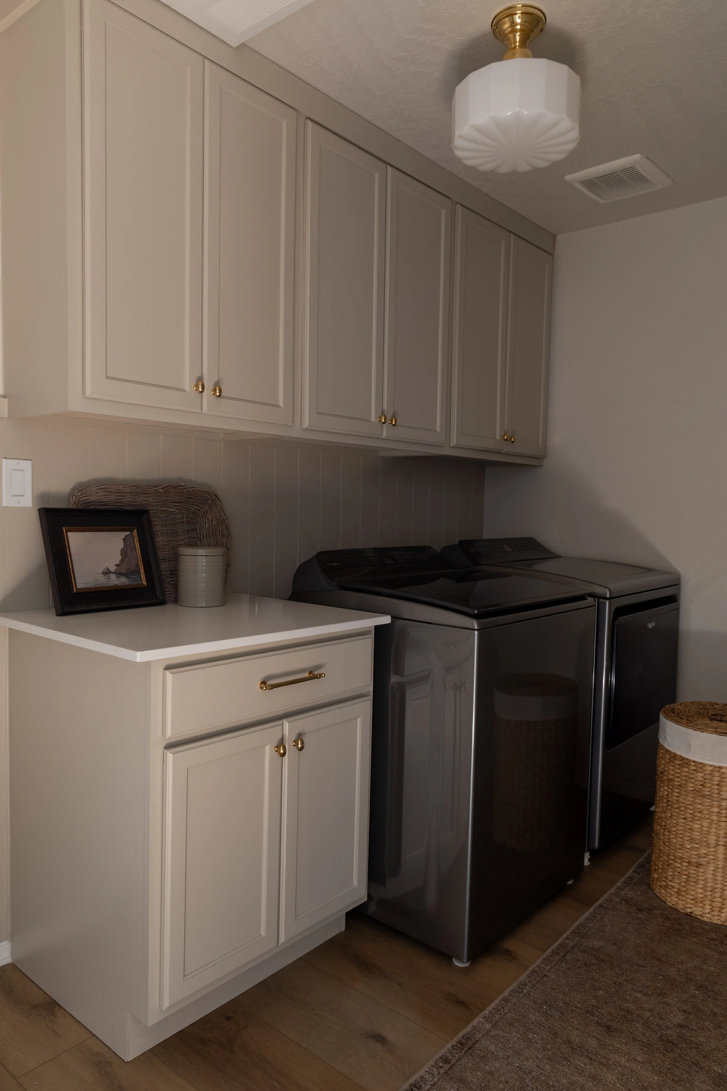A luxury laundry room with white cabinets, a white countertop, a framed picture, a ribbed basket, a white vase, and a top-loading washer and dryer in stainless steel. There is a wicker laundry basket and a decorative basket on a rug.