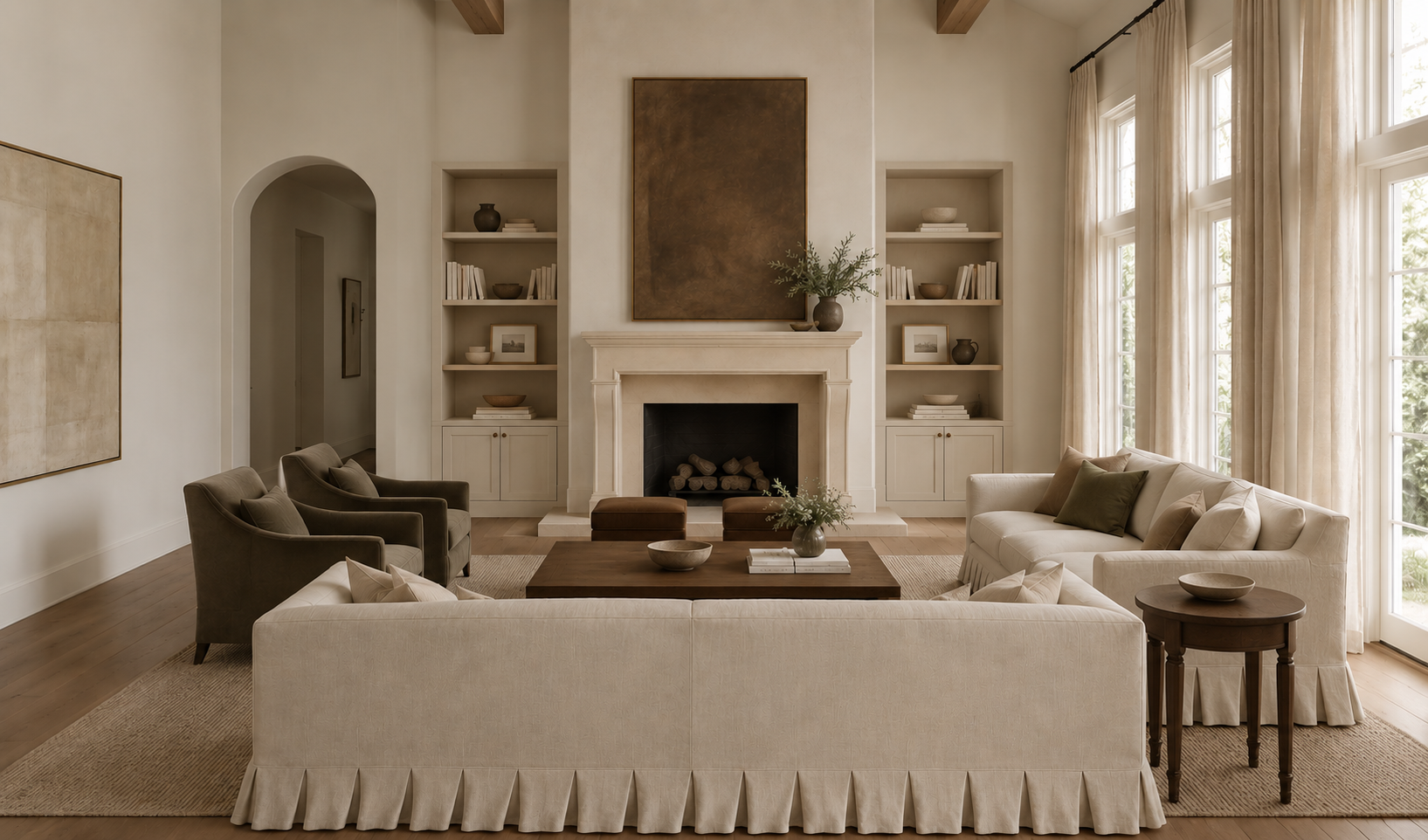 Living room with white walls, large windows with curtains, beige sofas, armchairs, a wooden coffee table, a fireplace with a brown painting above, and built-in shelves with books and decorative objects.
