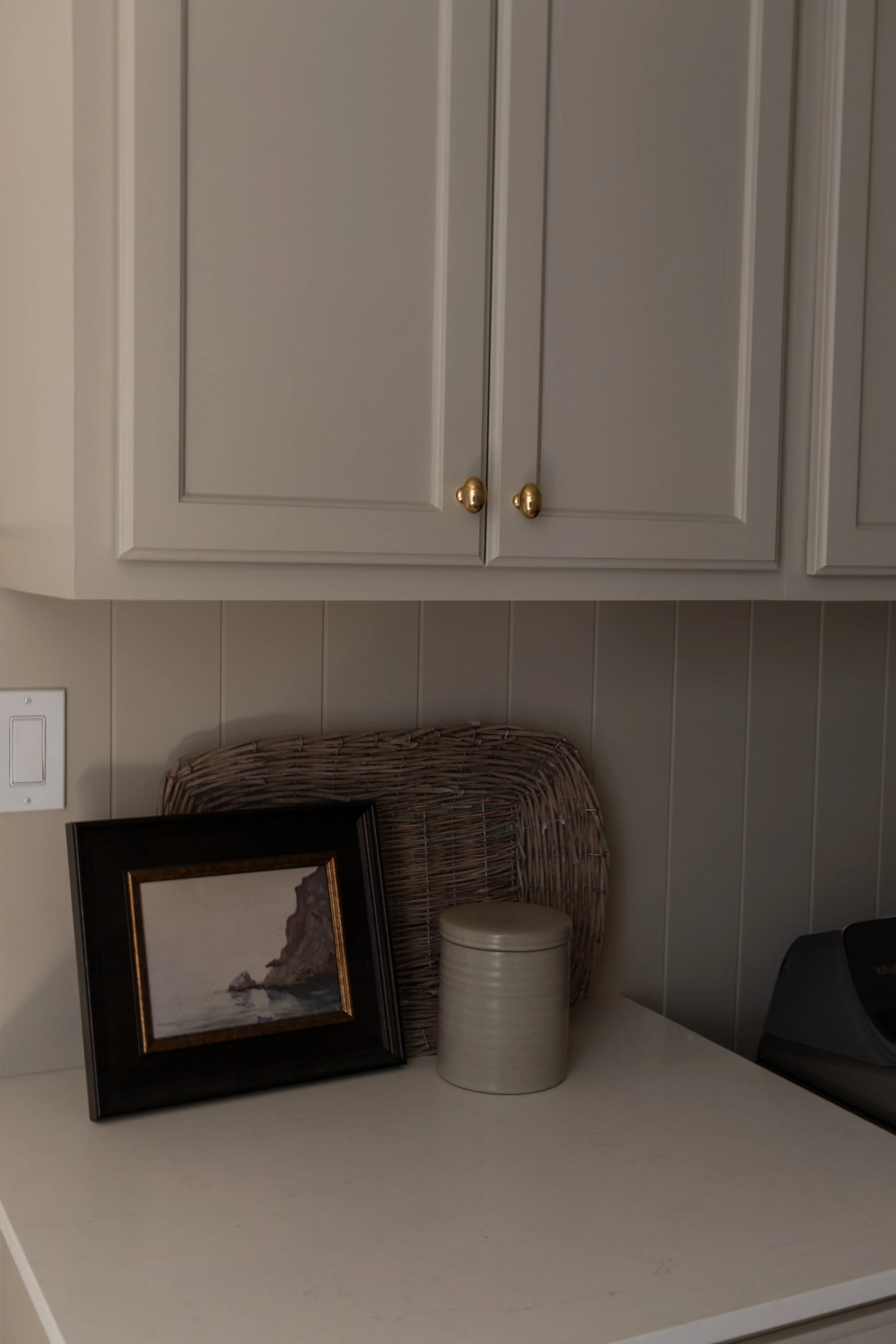 Laundry roomwith Drop Cloth cabinet color with rounded brass knobs, panelled wall, framed mountain landscape photo, wicker basket, beige ceramic container, and washer and dryer.