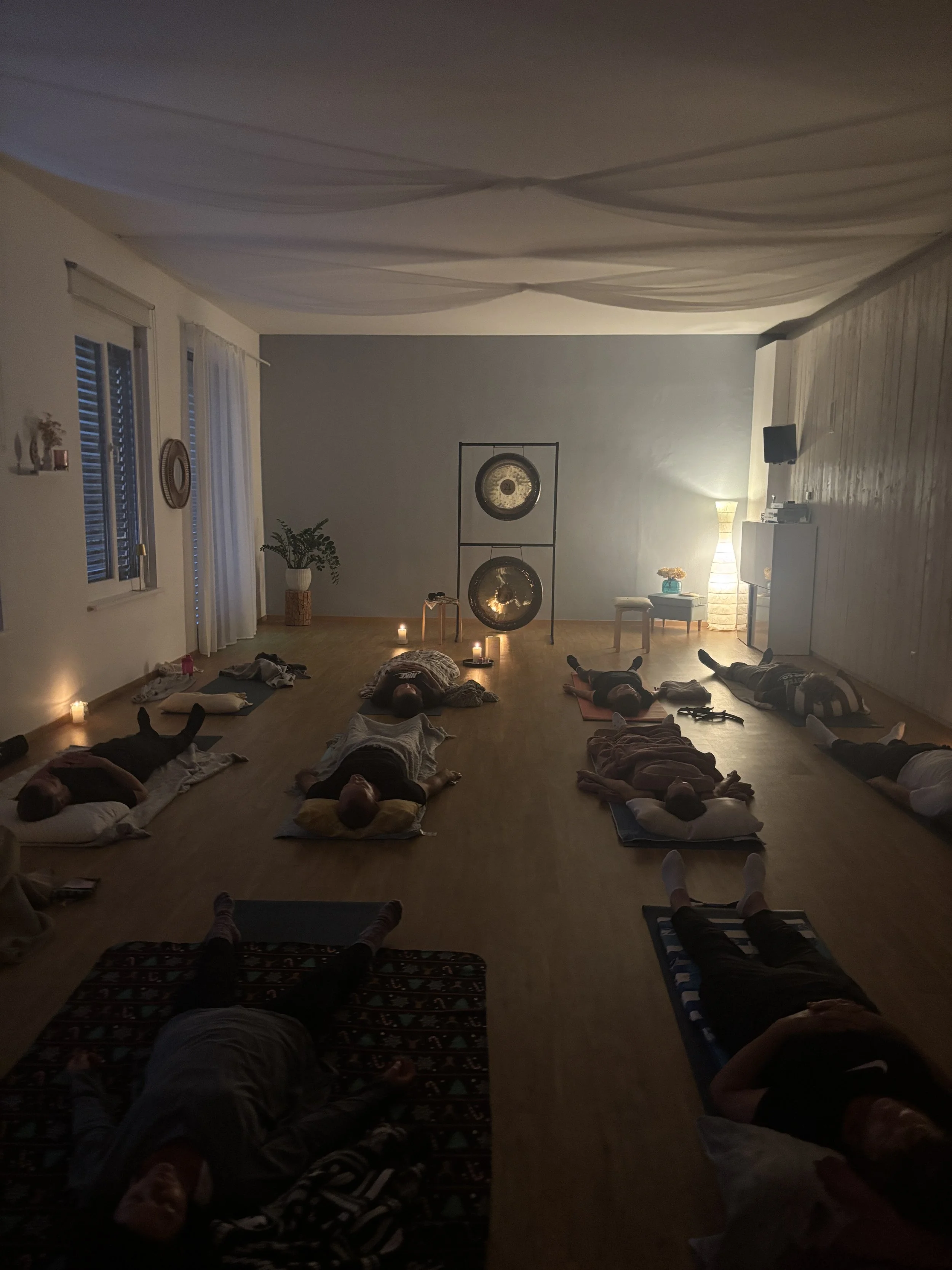Menschen liegen auf Yogamatten in einem mit Kerzenlicht erhellten  Raum, der für ein Gongbad vorbereitet ist, mit Gongs im Hintergrund.
