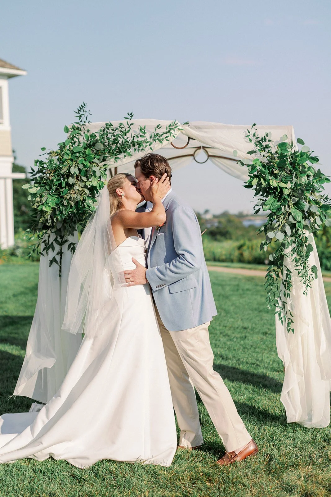 A bride and groom share a kiss beneath a wedding arch decorated with greenery and white fabric outdoors on a sunny day.