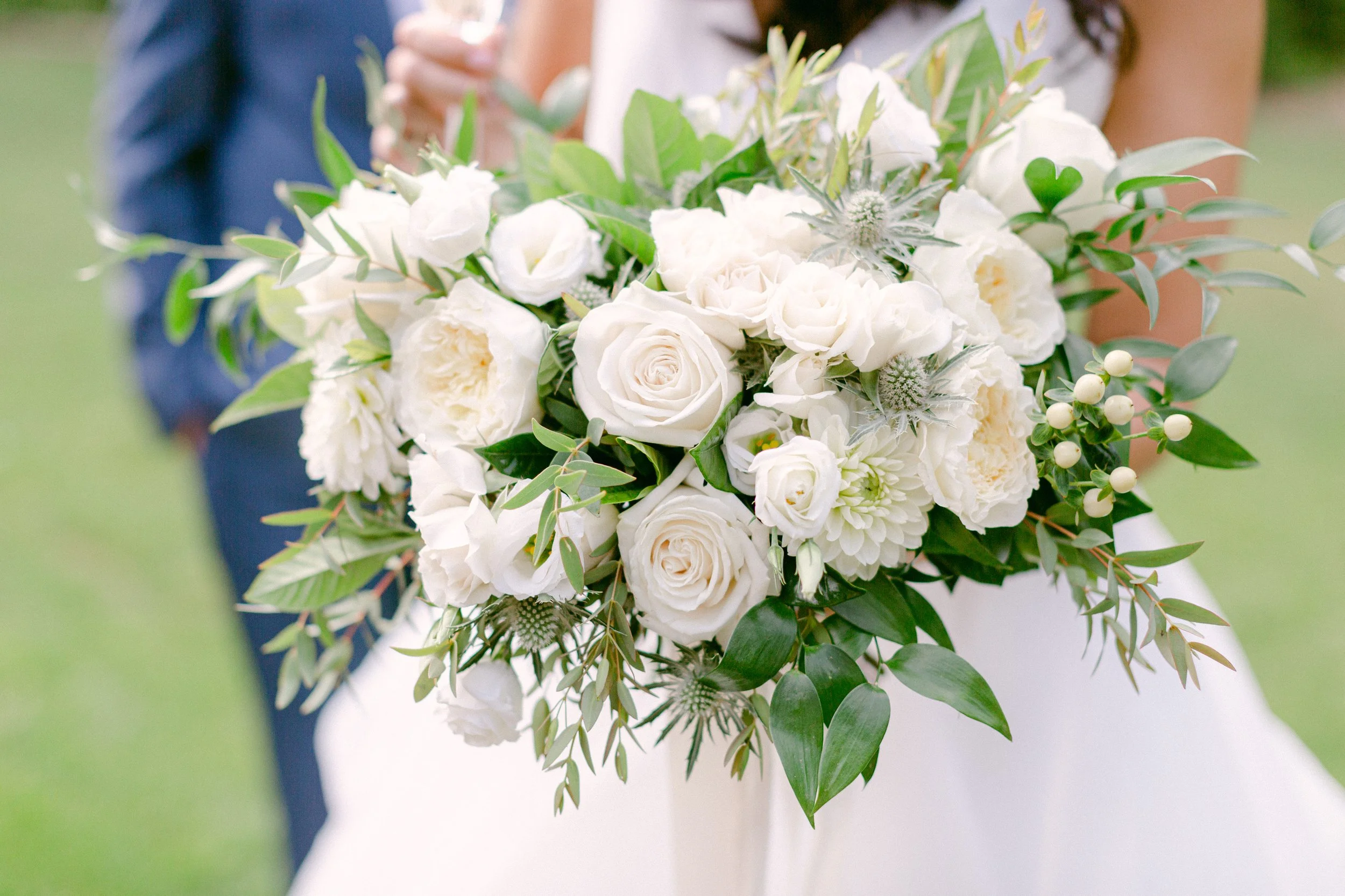 A bride holding a bouquet of white roses, dahlias, and eucalyptus leaves at an outdoor wedding.