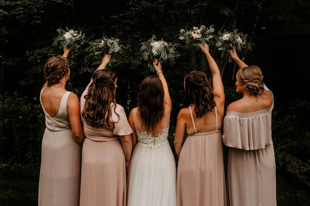 Five women in light-colored dresses stand with their backs to the camera, holding floral bouquets high above their heads in a wooded area.