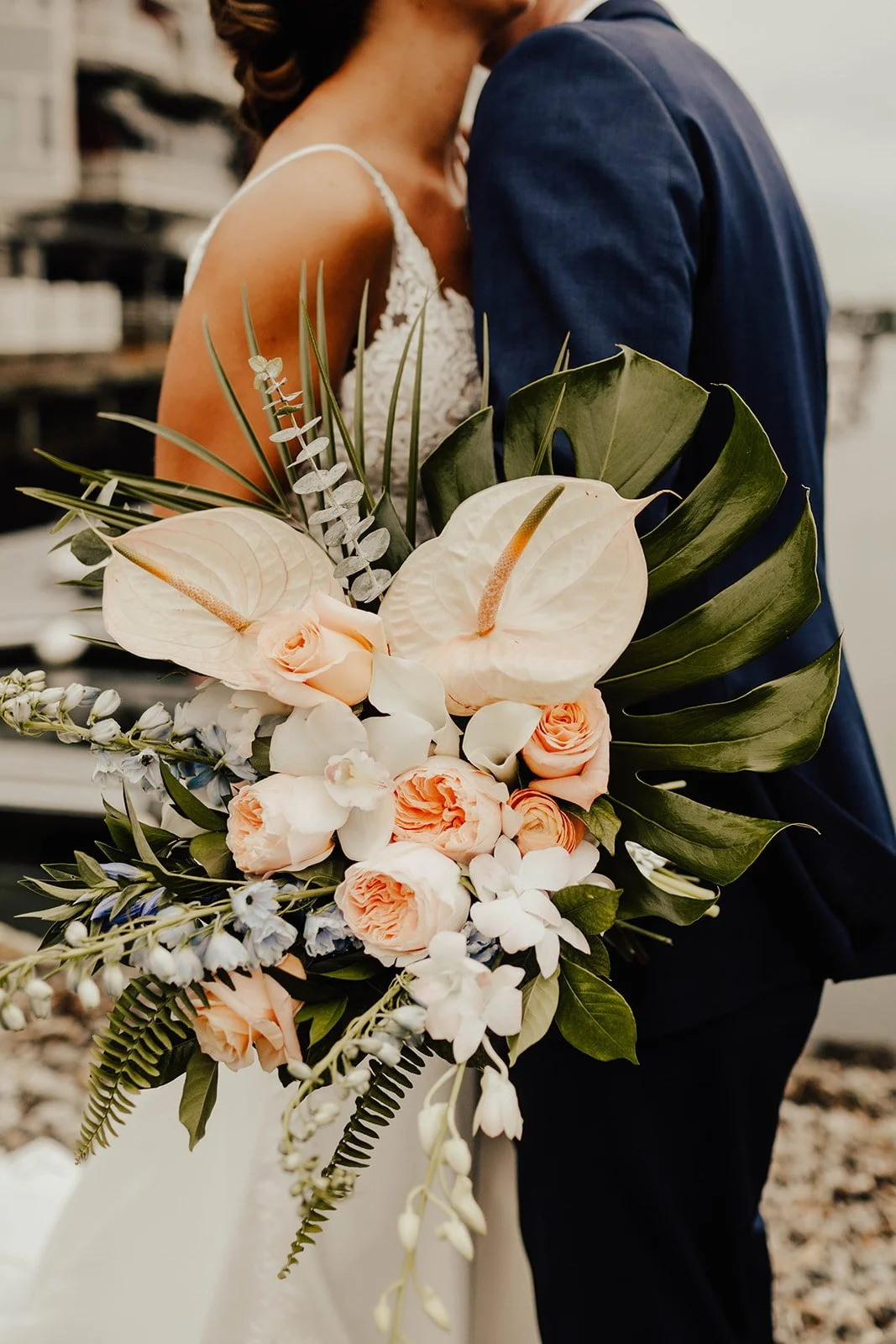 Close-up of a bride and groom holding a large bouquet of pastel pink roses, white orchids, and greenery, with the bride in a lace dress and the groom in a navy suit.