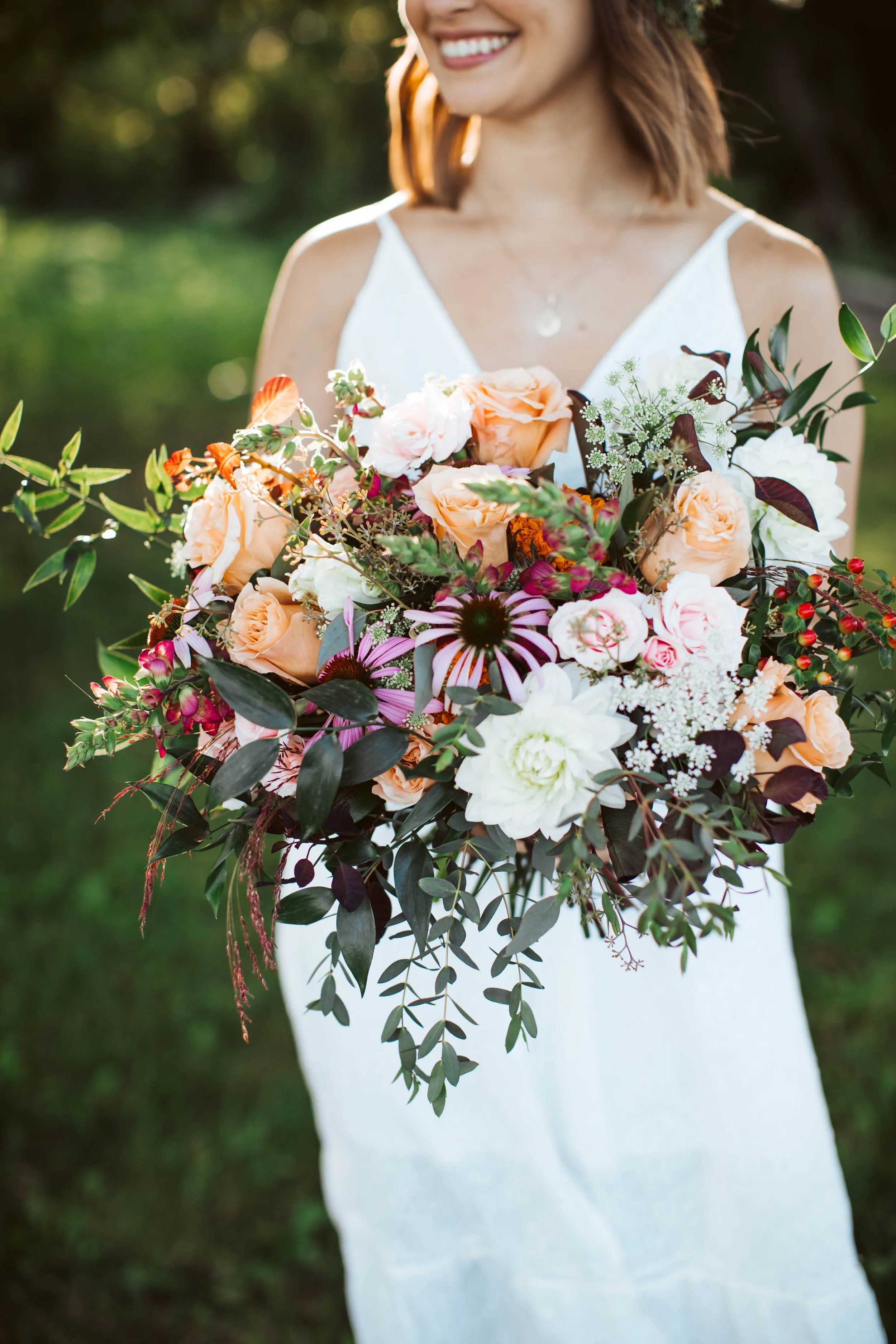 A woman in a white dress holding a large colorful flower bouquet outdoors, smiling with a blurred green background.
