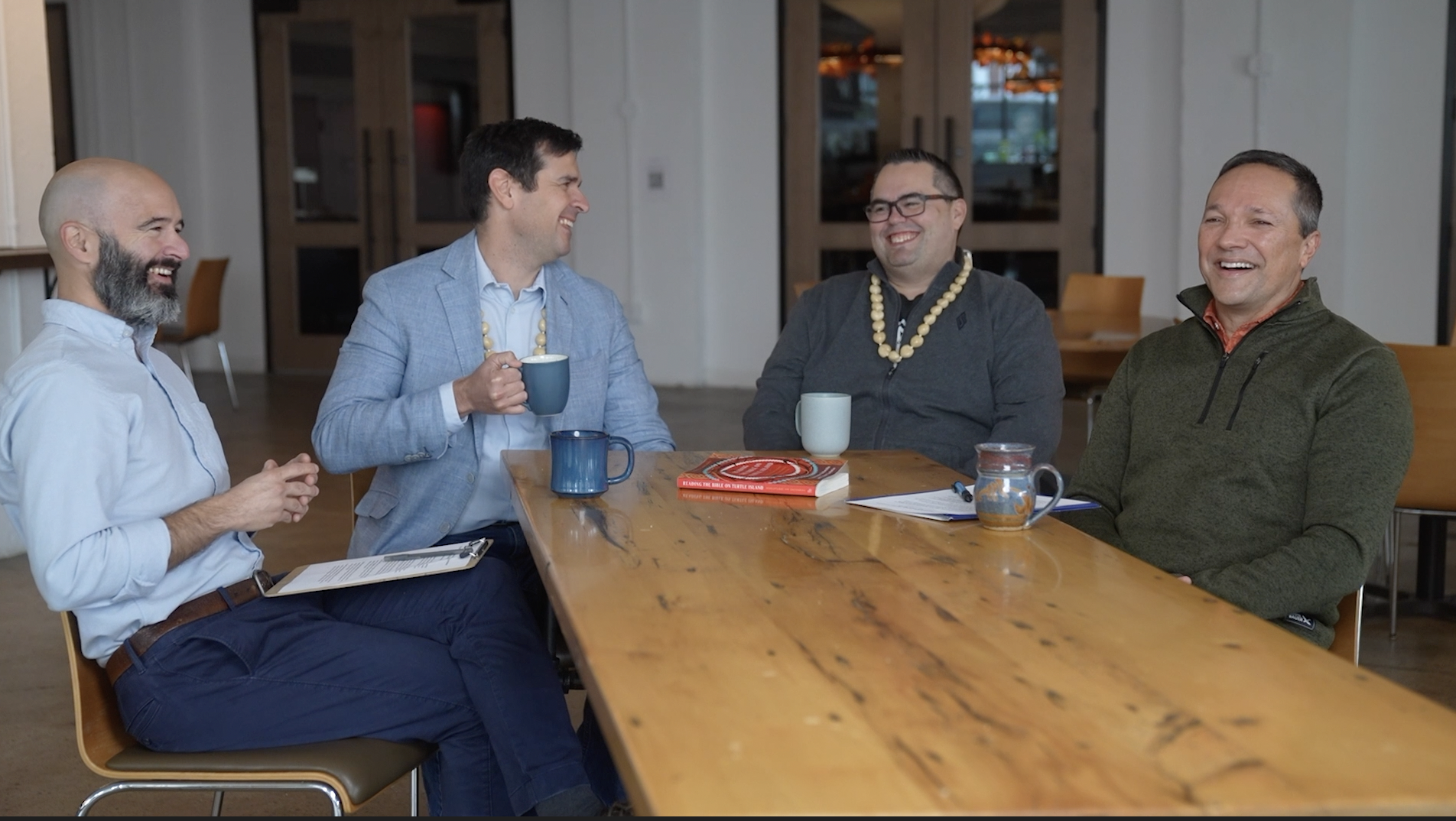 Four men sit around a wooden table drinking coffee and laughing