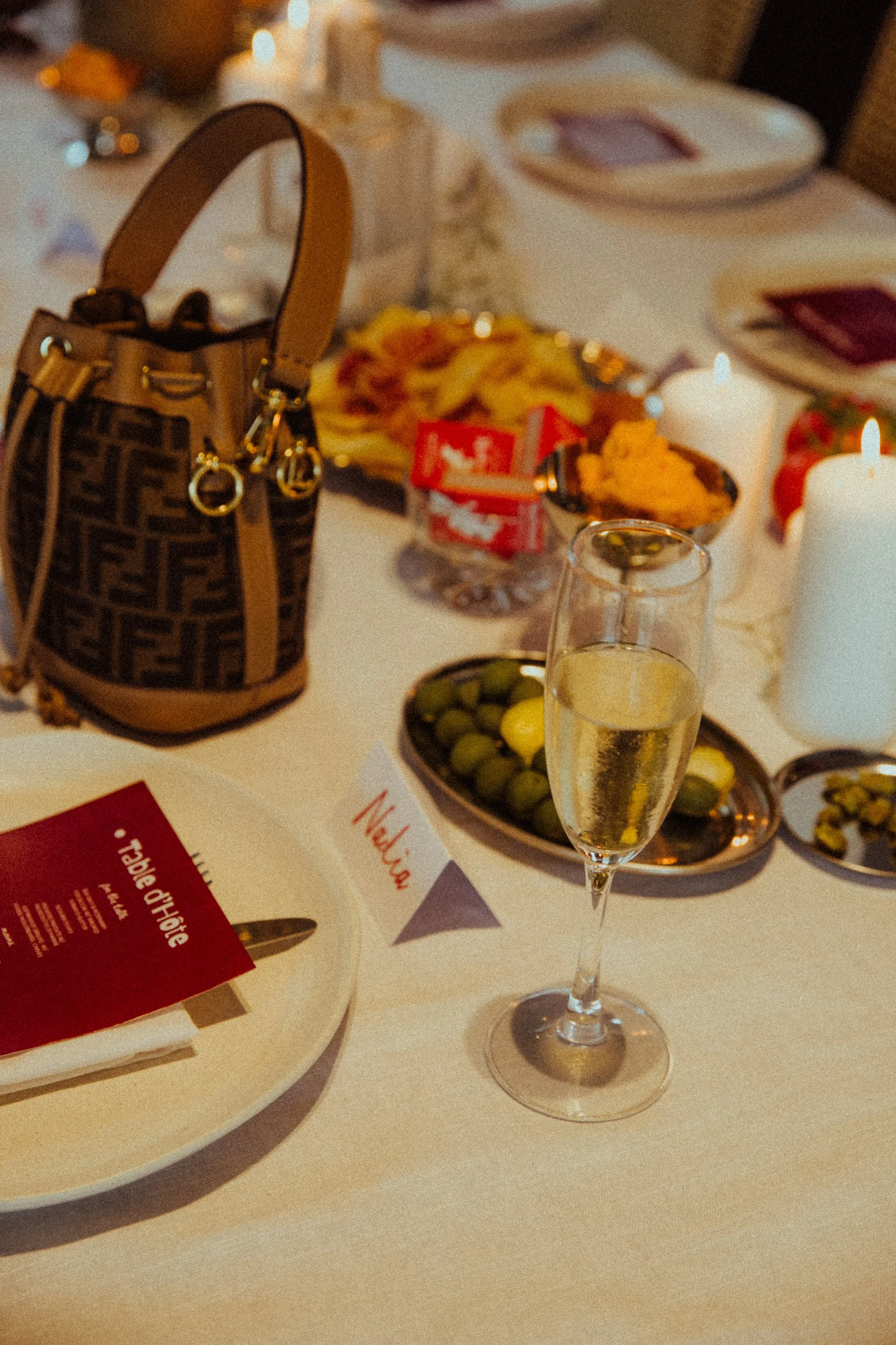 A dinner table set with plates, a glass of champagne, candles, and a purse. There are various dishes including olives, chips, and other appetizers, with a name card that reads "Nelvia".