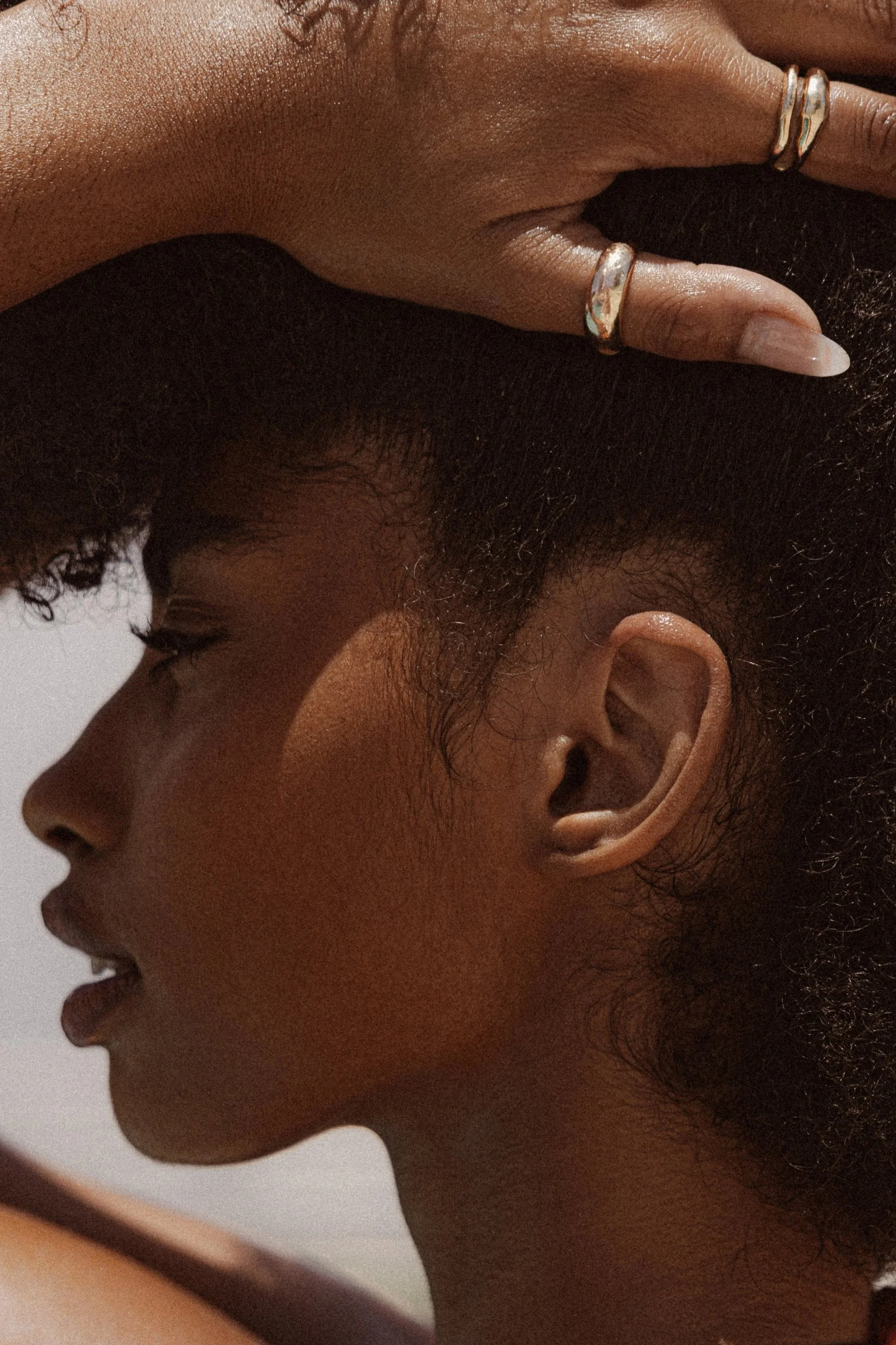 Close-up side profile of a woman with natural hair, hands resting on her head, wearing rings on her fingers.
