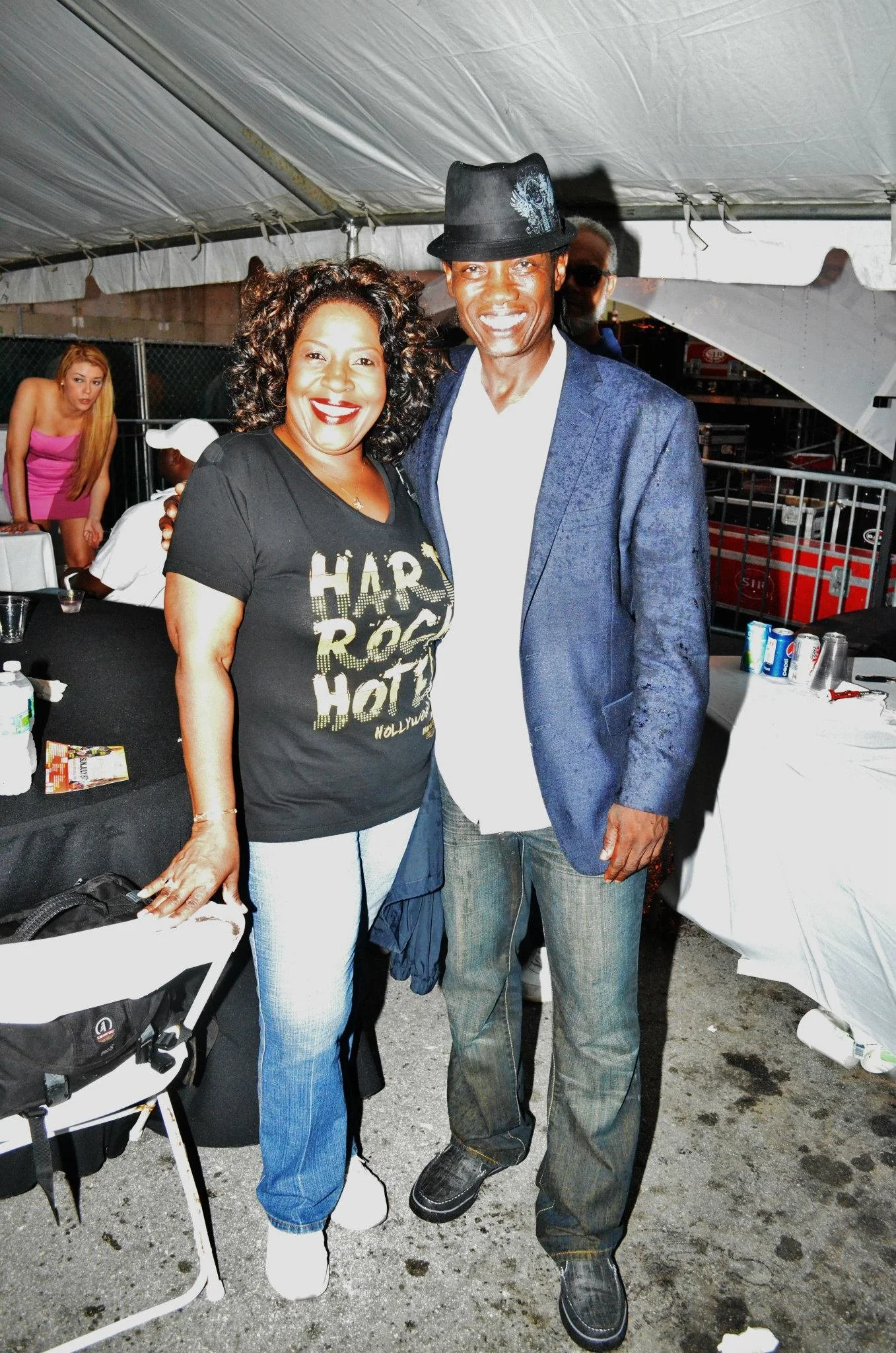 A woman and a man pose together under a tent at an outdoor event, smiling at the camera. The woman wears a black T-shirt with gold text, and the man wears a dark hat, a blue blazer, and jeans.