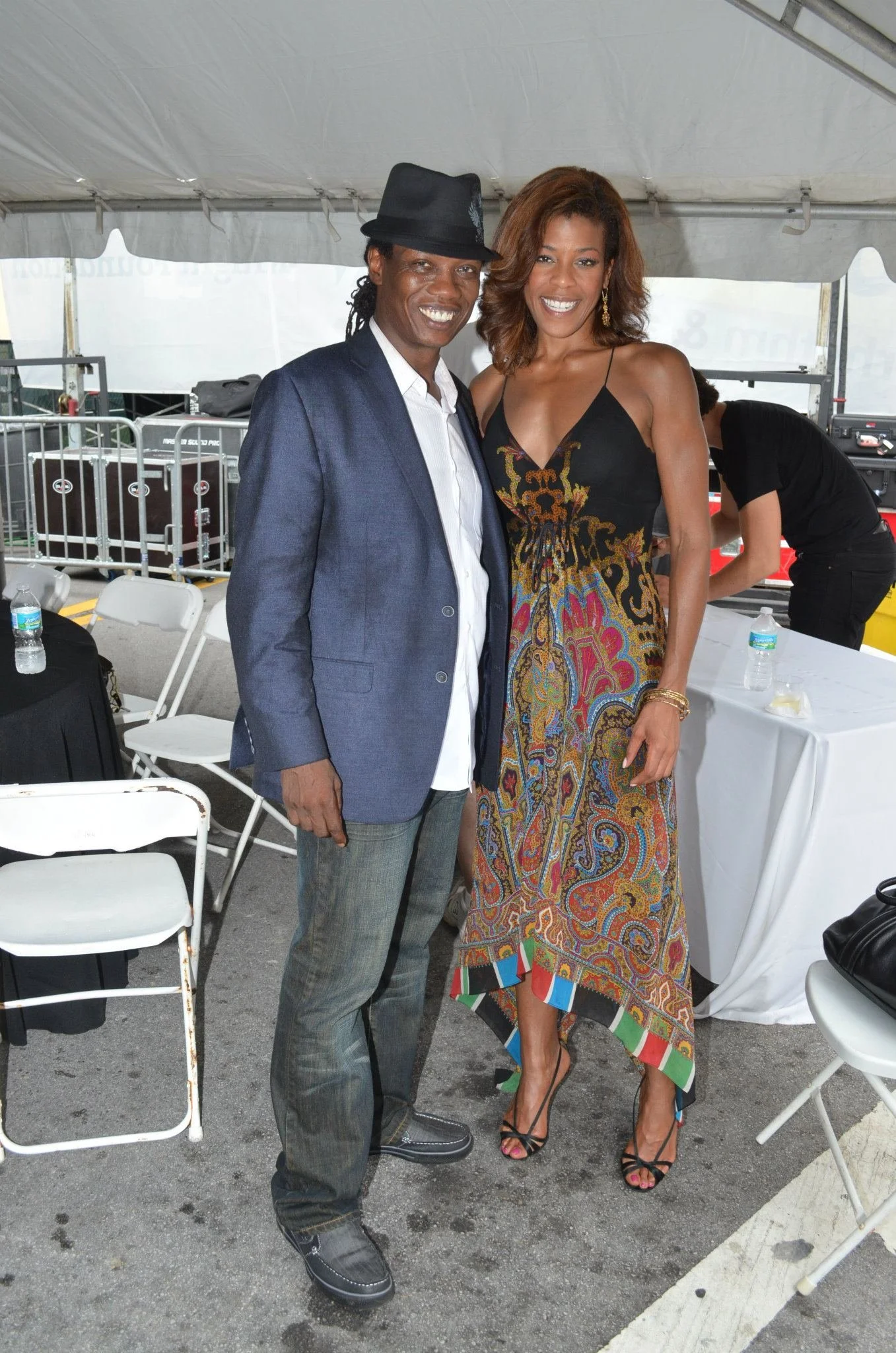 A man and woman standing together under a tent, posing for a photo. The man is wearing a dark hat, white shirt, blue blazer, and gray pants. The woman is wearing a colorful patterned dress with thin straps and black high-heeled sandals. They are smil