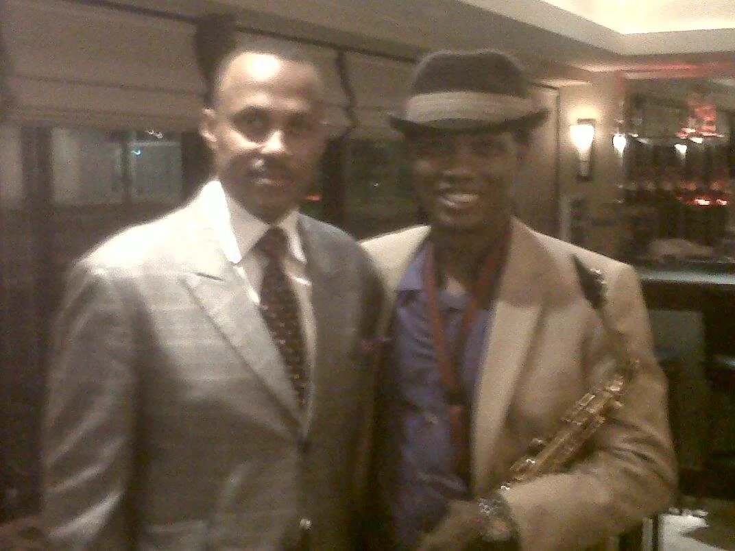Two men in suits posing together in a dimly lit indoor setting. One is holding a saxophone.