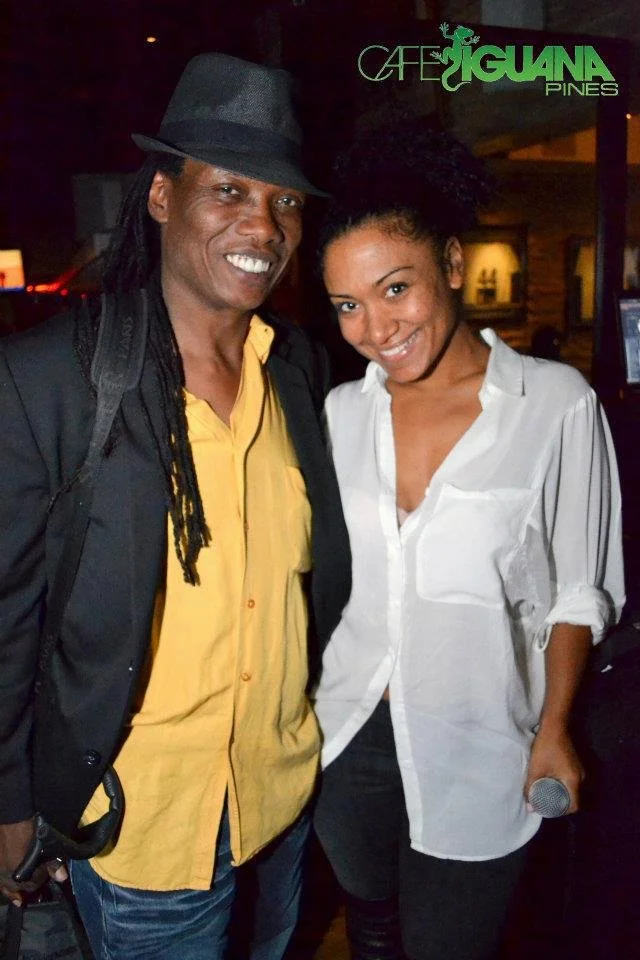 Two smiling people, a man with long dreadlocks wearing a black hat and black blazer over a yellow shirt, and a woman in a white shirt holding a microphone, standing inside Cafe Iguana Pines.