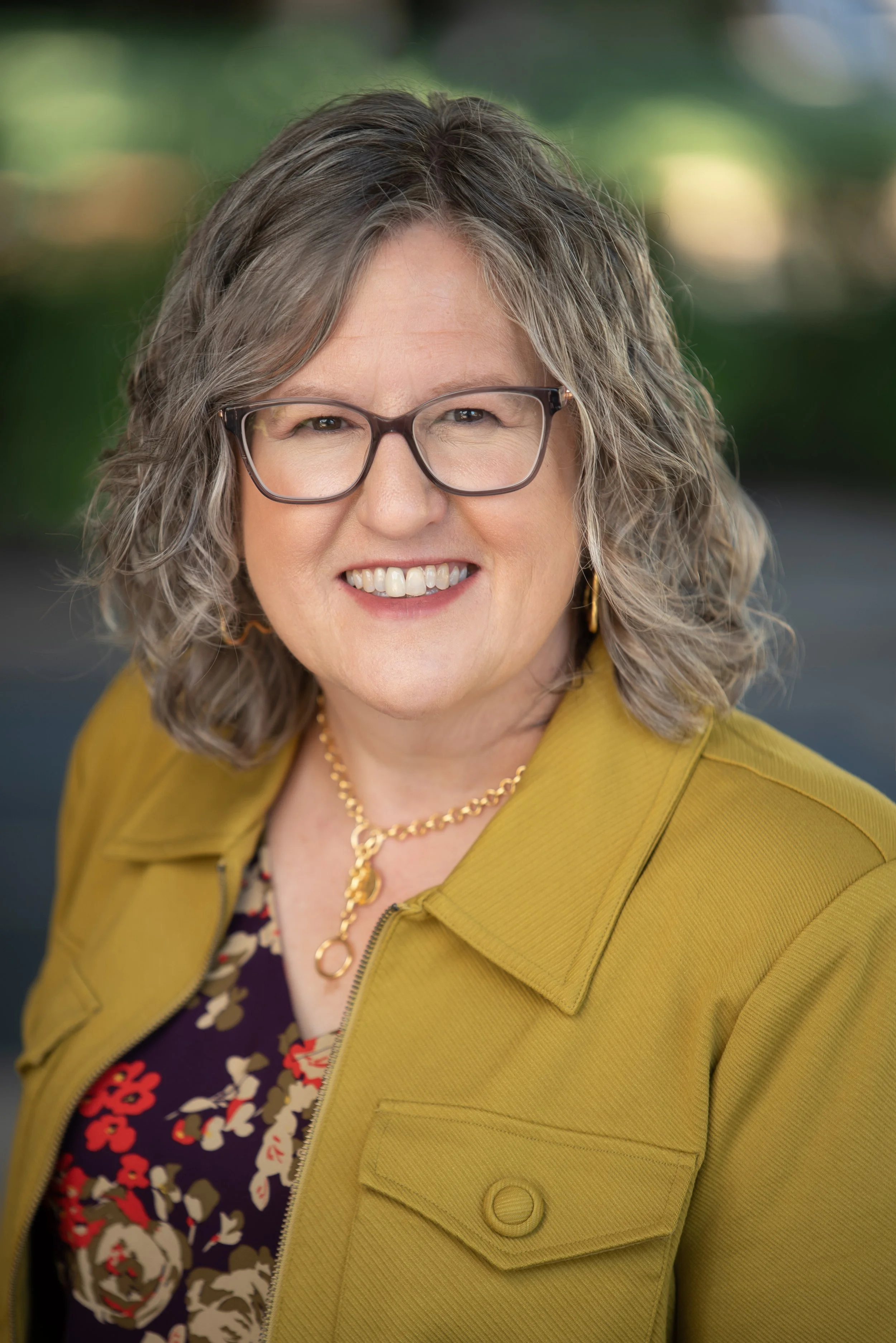 A smiling woman with curly gray hair wearing glasses, a mustard yellow jacket, a floral blouse, a gold necklace, and earrings, outdoors with a blurred green background.