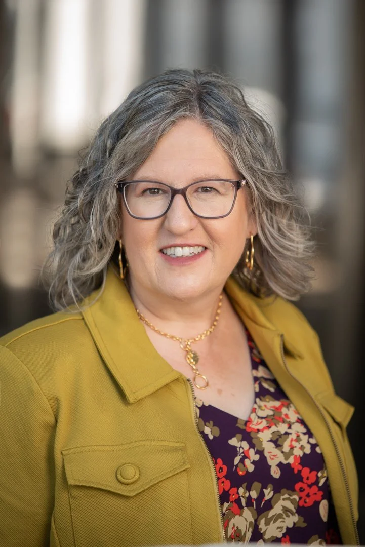 A woman with gray curly hair, wearing glasses, a gold necklace, gold hoop earrings, a yellow jacket, and a floral top, smiling at the camera.