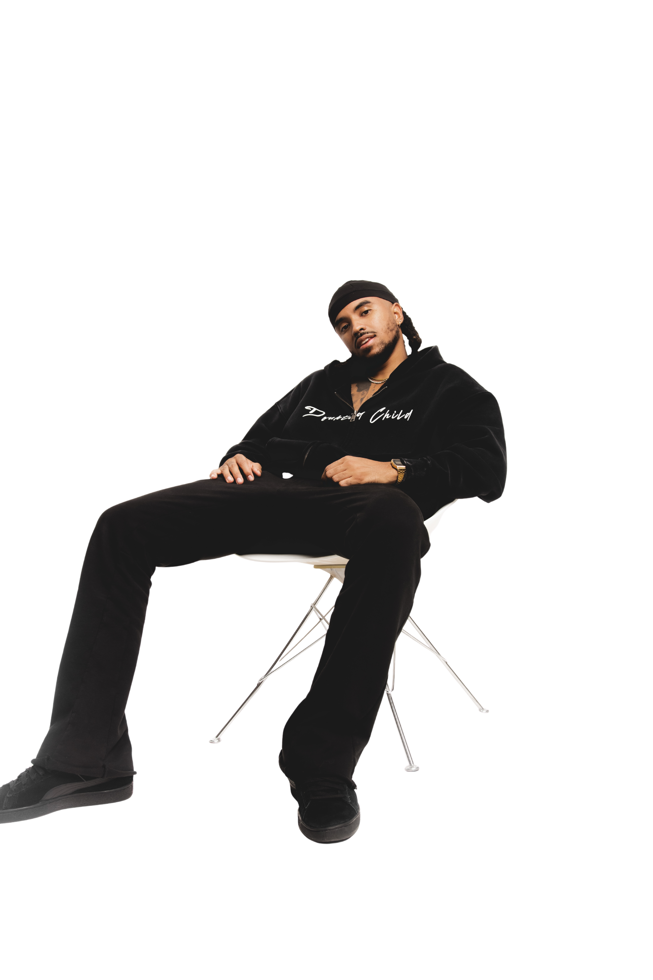 A young man sitting casually on a modern white chair with metal legs, wearing a black hoodie, black pants, a black wave cap, a gold watch, and a gold chain, with a relaxed and confident expression.