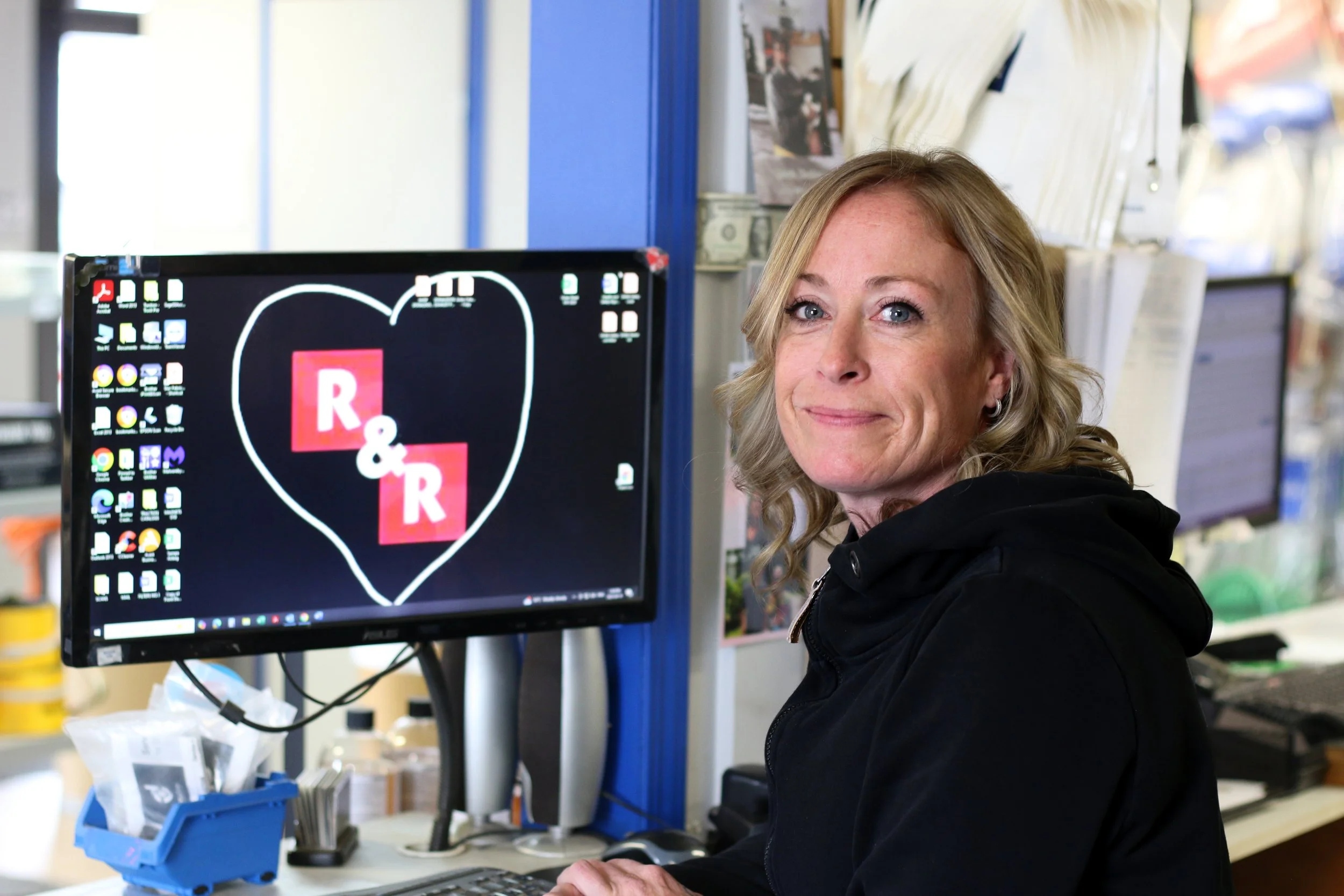 A woman with blonde curly hair sitting at a desk in front of a computer monitor with a black background and a heart outline surrounding red squares with white letters "R& R." She is smiling and wearing a black hoodie.