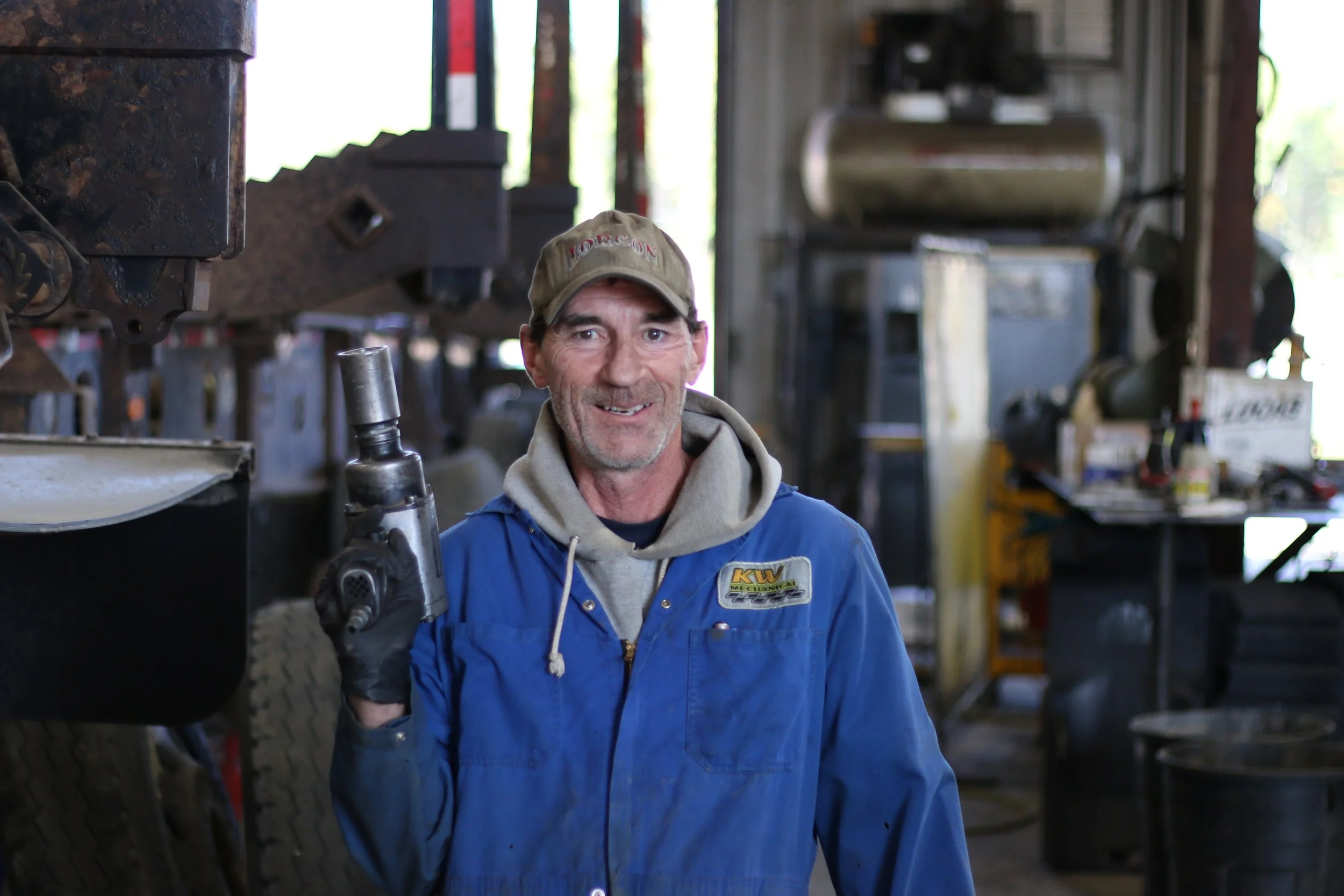 A smiling man with a beard wearing a blue mechanic's jacket, a beige hoodie, a cap, and black gloves holding a cordless power drill inside a workshop with industrial equipment.