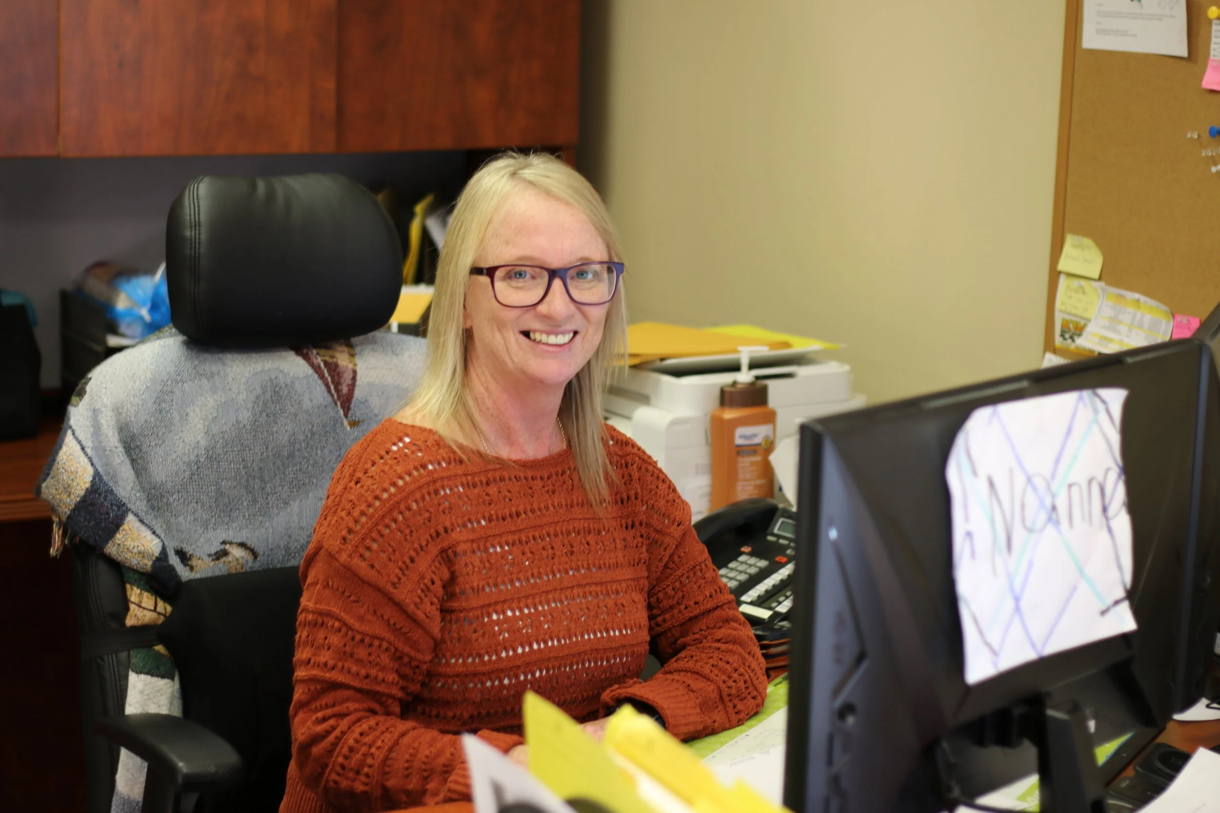 A woman with blonde hair, glasses, and a warm smile sitting at a desk in an office. She is wearing a rust-colored knit sweater, surrounded by office supplies and a computer monitor.