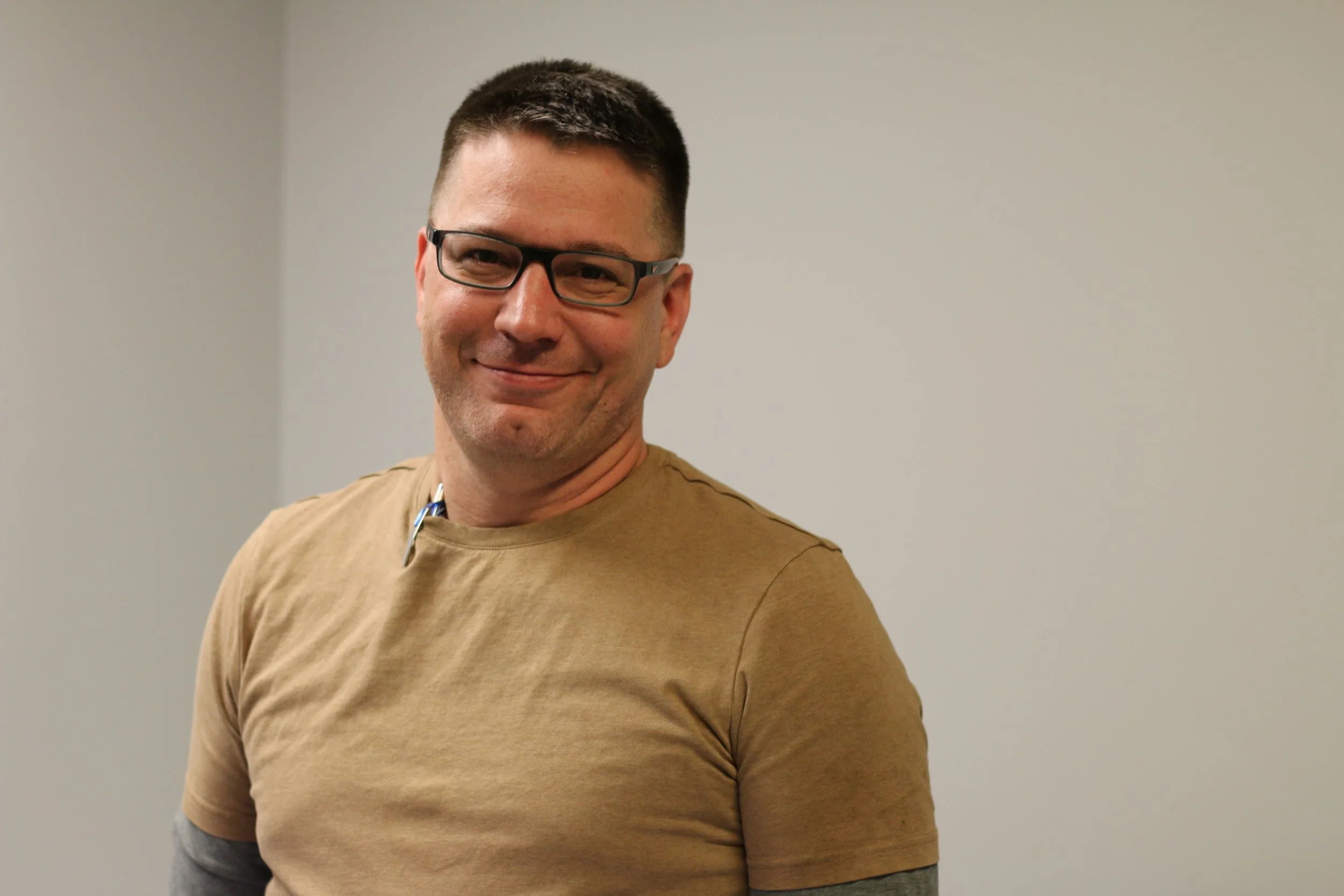 A man with short dark hair, glasses, and a light brown t-shirt smiling in front of a plain wall.