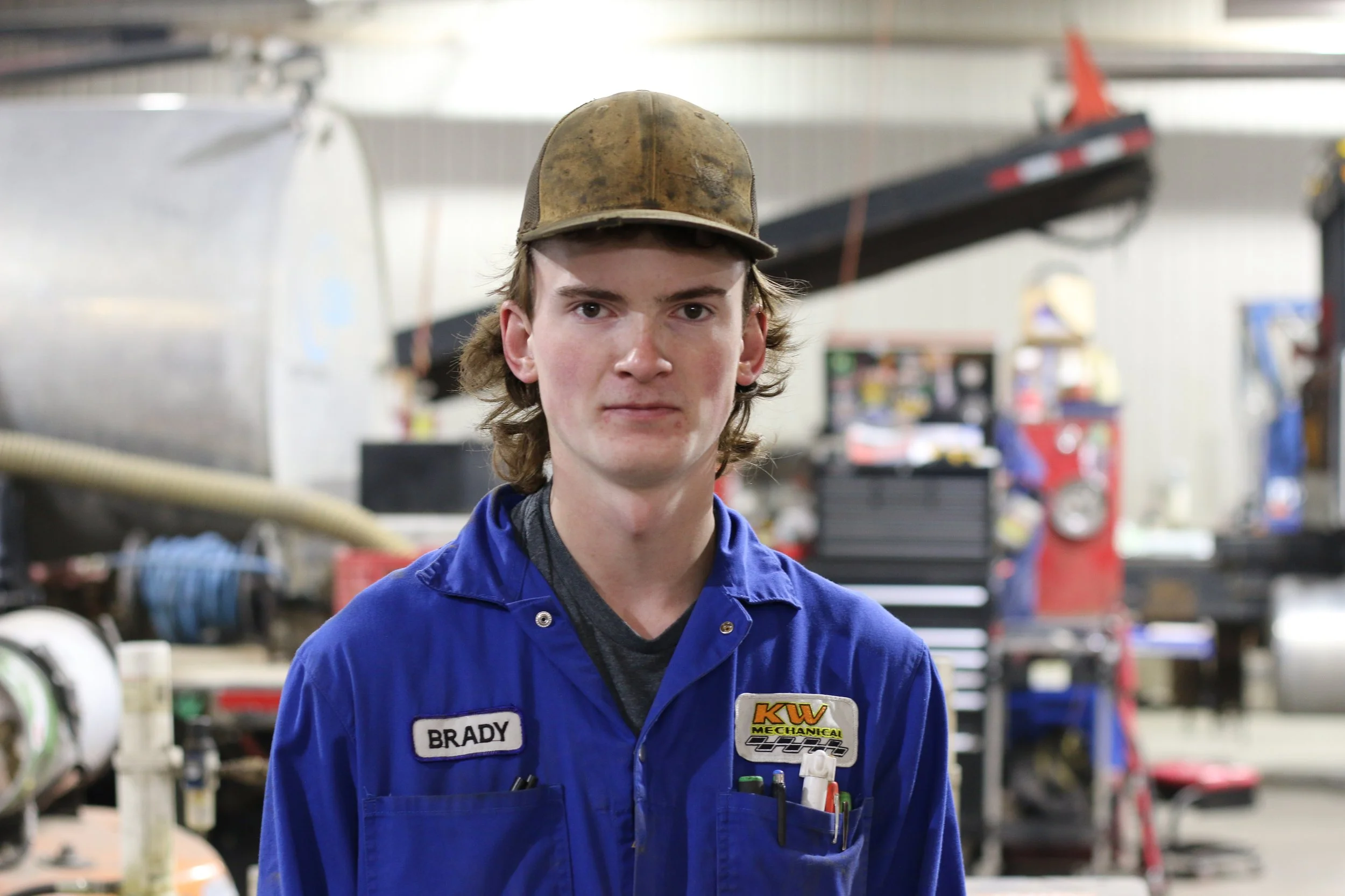 A young man in a blue mechanic's uniform with the name tag 'Brady' stands in a workshop. He wears a brown cap, and behind him are tools and equipment, including a red and black crane and various workbenches.