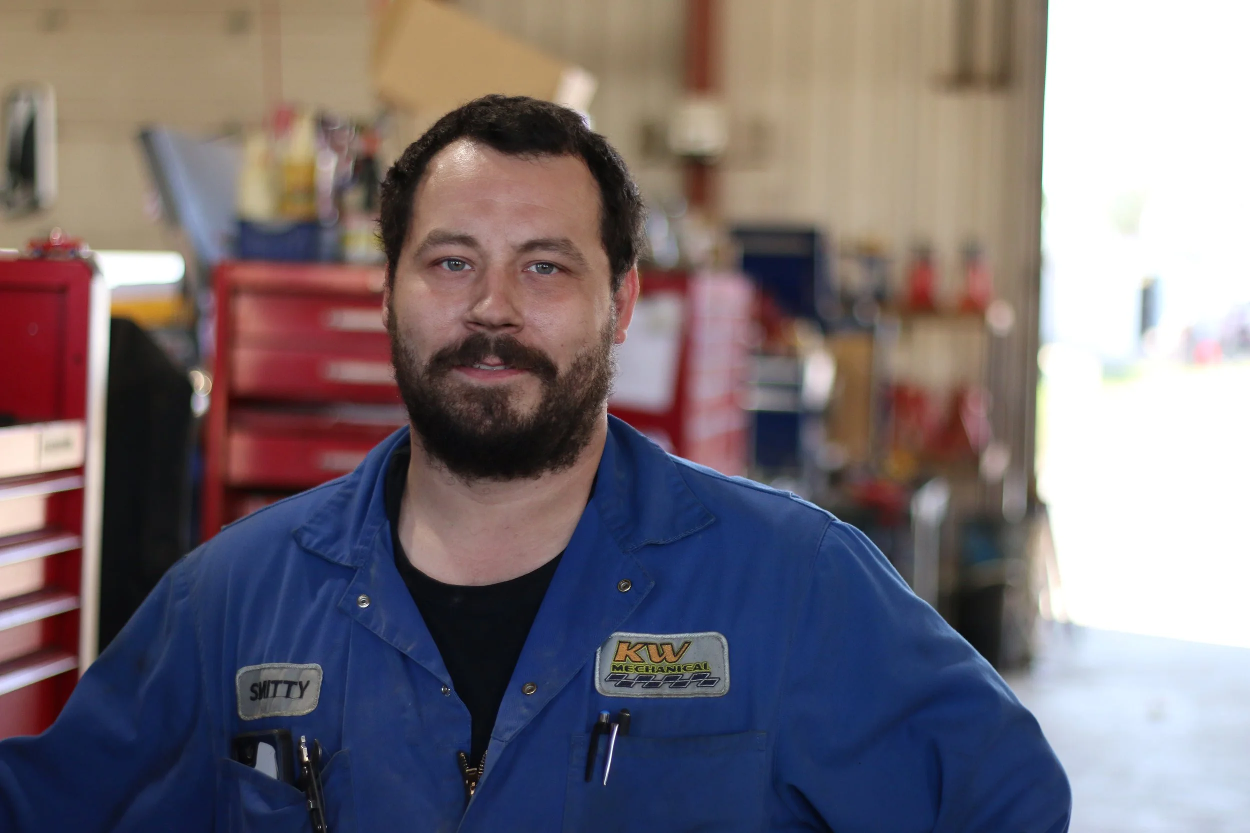 A mechanic with dark hair and a beard wearing a blue work jumpsuit standing inside a garage, with red tool chests and workshop equipment in the background.