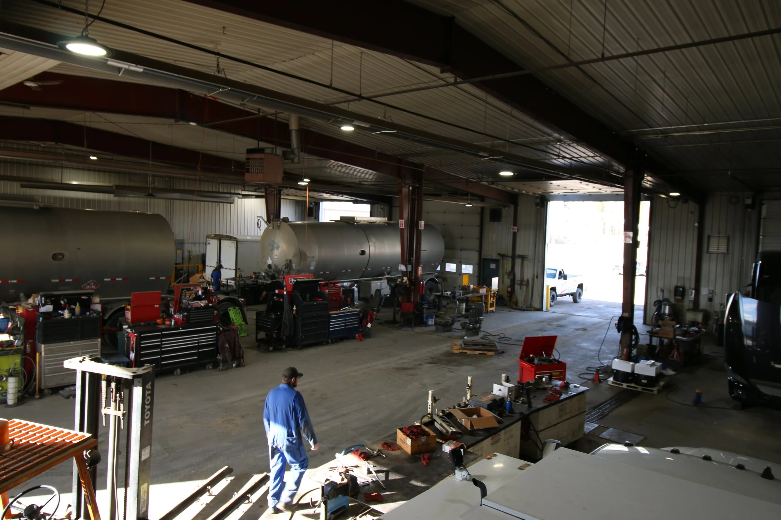 Inside an industrial workshop with two large metal tanks, various workstations, and a worker in blue overalls walking.