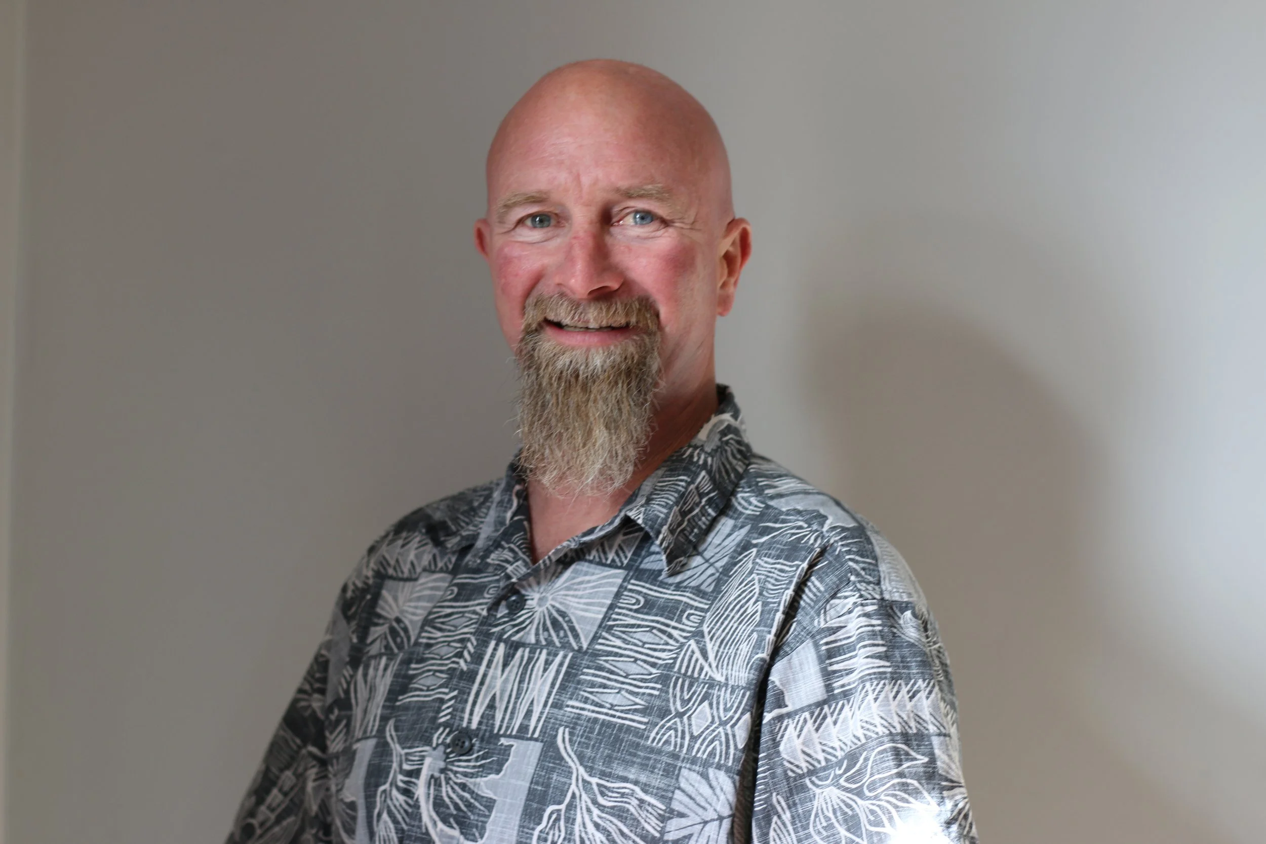 A smiling man with a bald head and a goatee, wearing a patterned button-up shirt, standing against a plain wall.