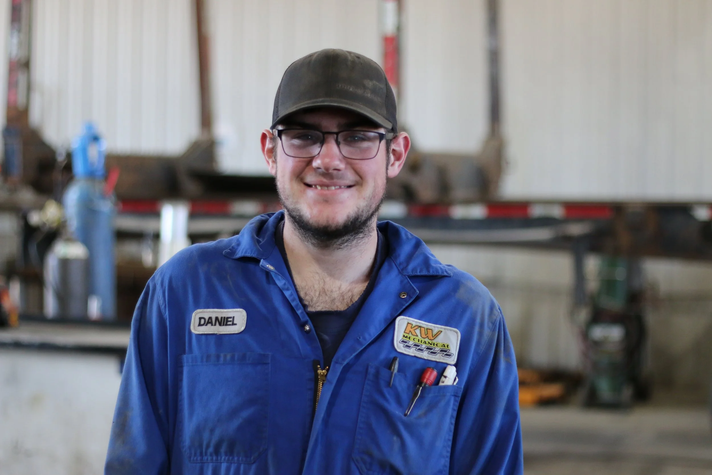 A smiling young man wearing glasses and a blue work uniform with a name tag that says Daniel, standing inside a workshop or garage.