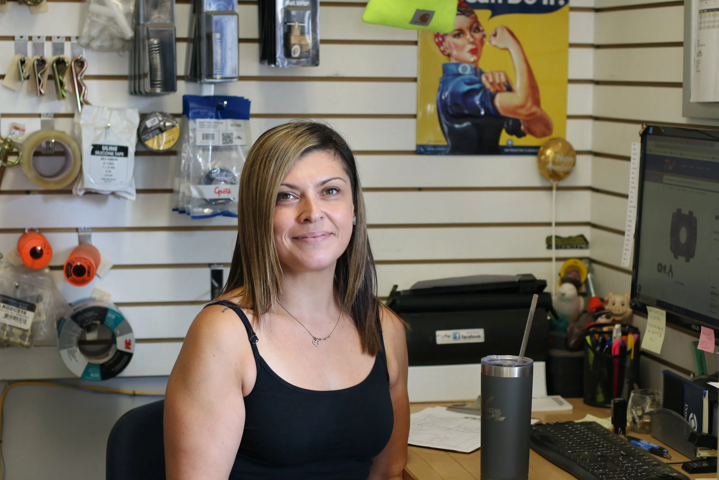 Woman sitting at a desk in a store or office, with computer and various tools and supplies on the wall behind her.